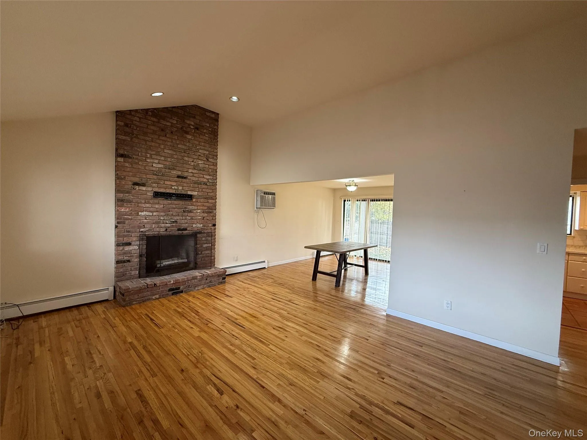 Unfurnished living room with a brick fireplace, hardwood / wood-style floors, a baseboard heating unit, high vaulted ceiling, and a wall mounted air conditioner Unfurnished living room with a brick fireplace, hardwood / wood-style floors, a baseboard heating unit, high vaulted ceiling, and a wall mounted air conditioner