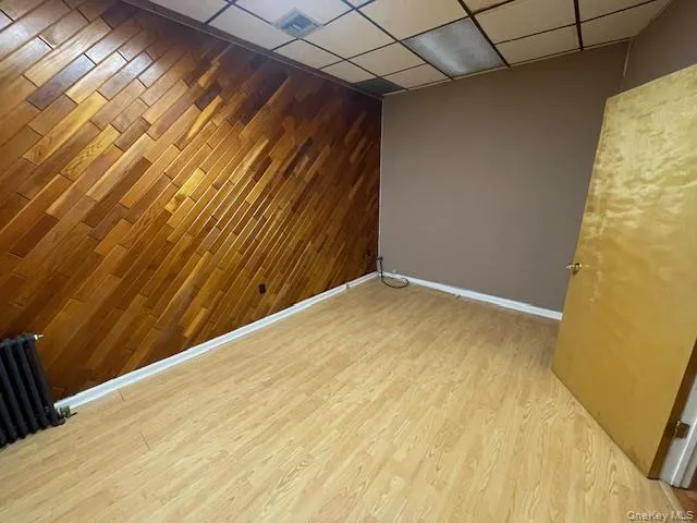 Empty room featuring wooden walls, light wood-style floors, an accent wall, radiator heating unit, and a drop ceiling Empty room featuring wooden walls, light wood-style floors, an accent wall, radiator heating unit, and a drop ceiling