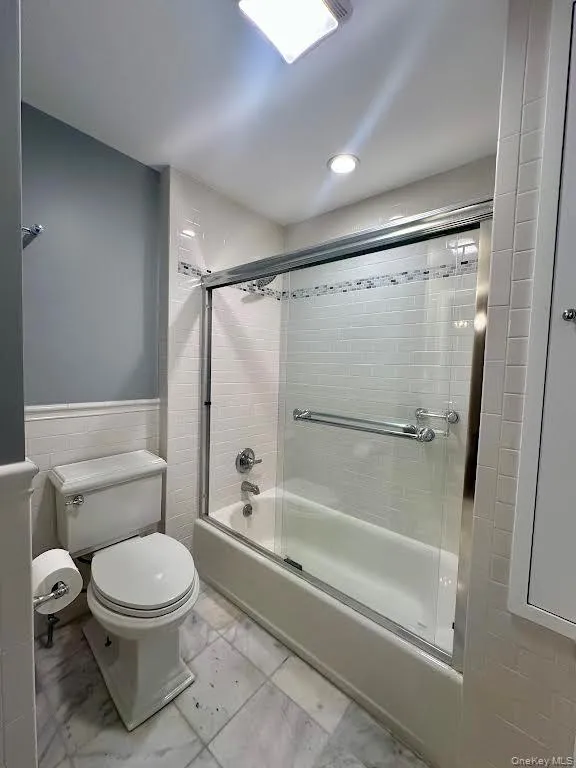 Bathroom with shower / bath combination with glass door, a wainscoted wall, and light marble finish flooring Bathroom with shower / bath combination with glass door, a wainscoted wall, and light marble finish flooring