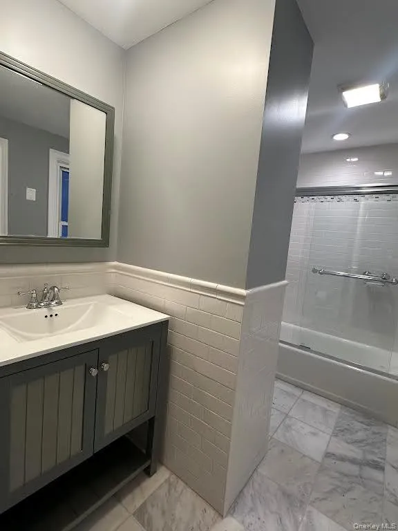 Bathroom featuring wainscoting, vanity, shower / bath combination with glass door, light marble finish flooring, and tile walls Bathroom featuring wainscoting, vanity, shower / bath combination with glass door, light marble finish flooring, and tile walls