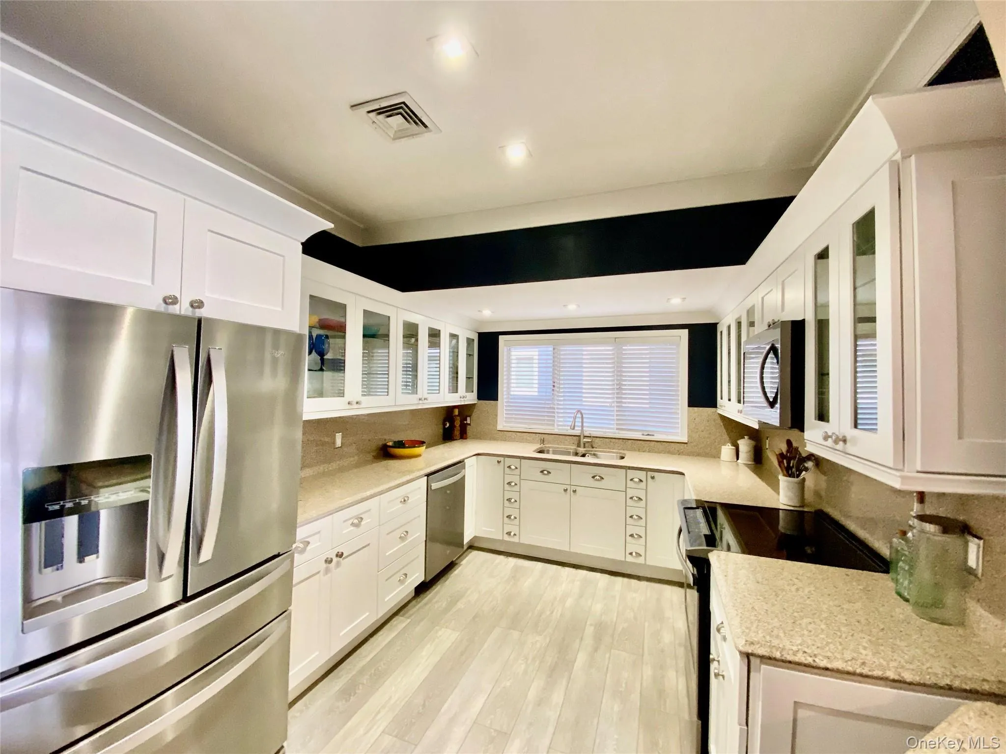 Kitchen with glass insert cabinets, appliances with stainless steel finishes, white cabinets, light stone countertops, and backsplash Kitchen with glass insert cabinets, appliances with stainless steel finishes, white cabinets, light stone countertops, and backsplash