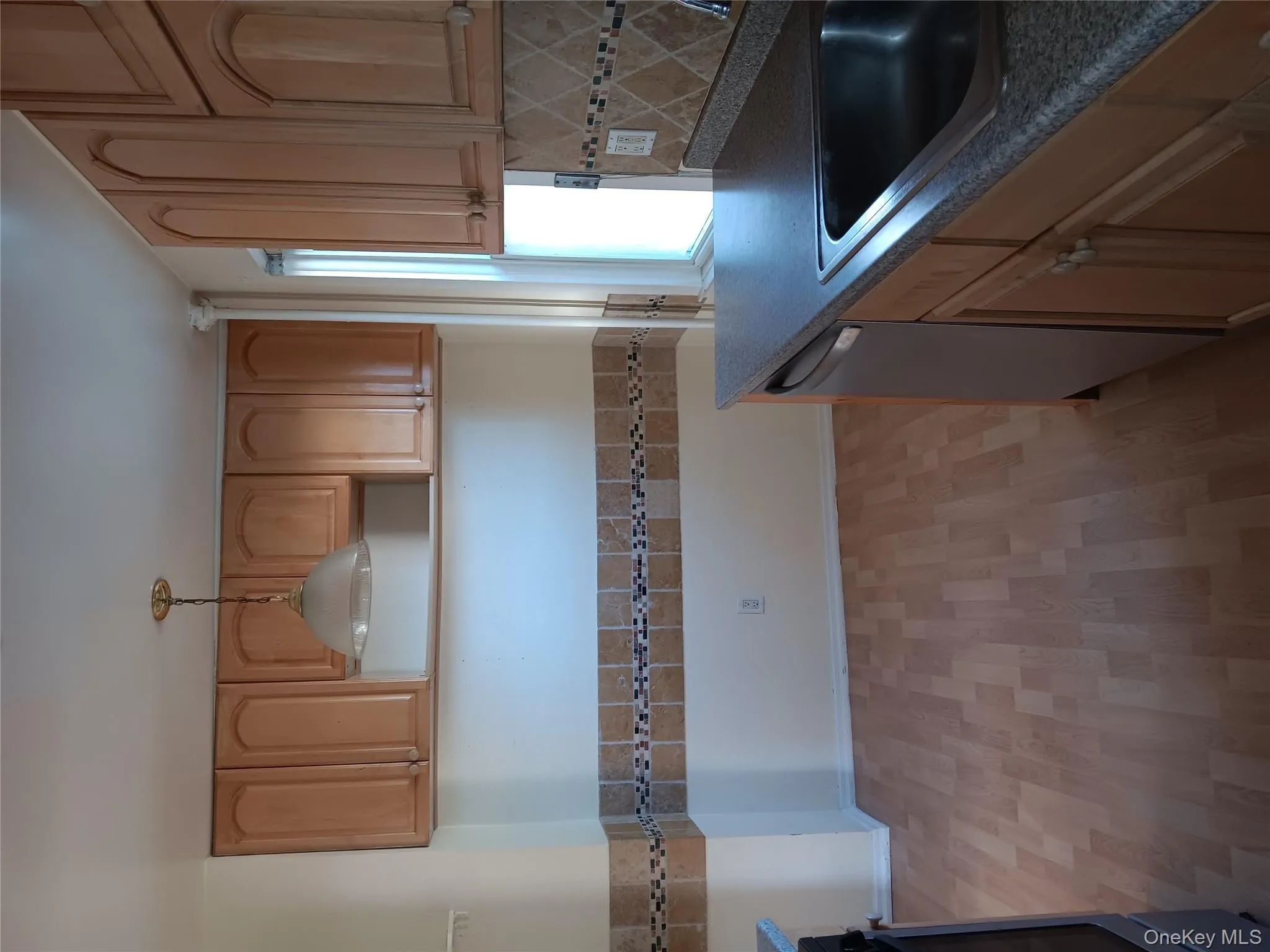 Kitchen with light wood-style flooring, dark countertops, dishwashing machine, hanging light fixtures, and backsplash Kitchen with light wood-style flooring, dark countertops, dishwashing machine, hanging light fixtures, and backsplash