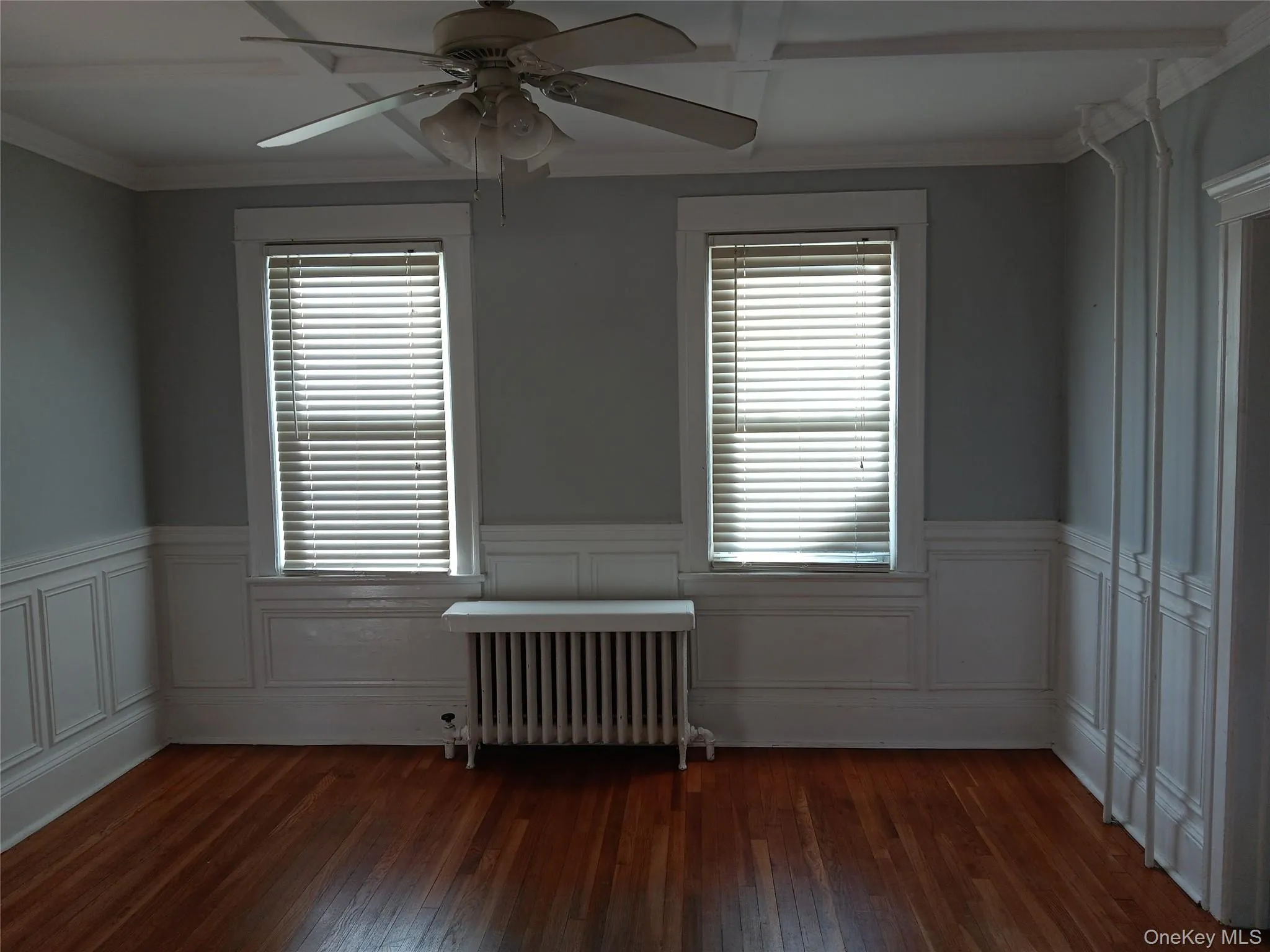 Empty room featuring ornamental molding, radiator, dark wood finished floors, a ceiling fan, and wainscoting Empty room featuring ornamental molding, radiator, dark wood finished floors, a ceiling fan, and wainscoting