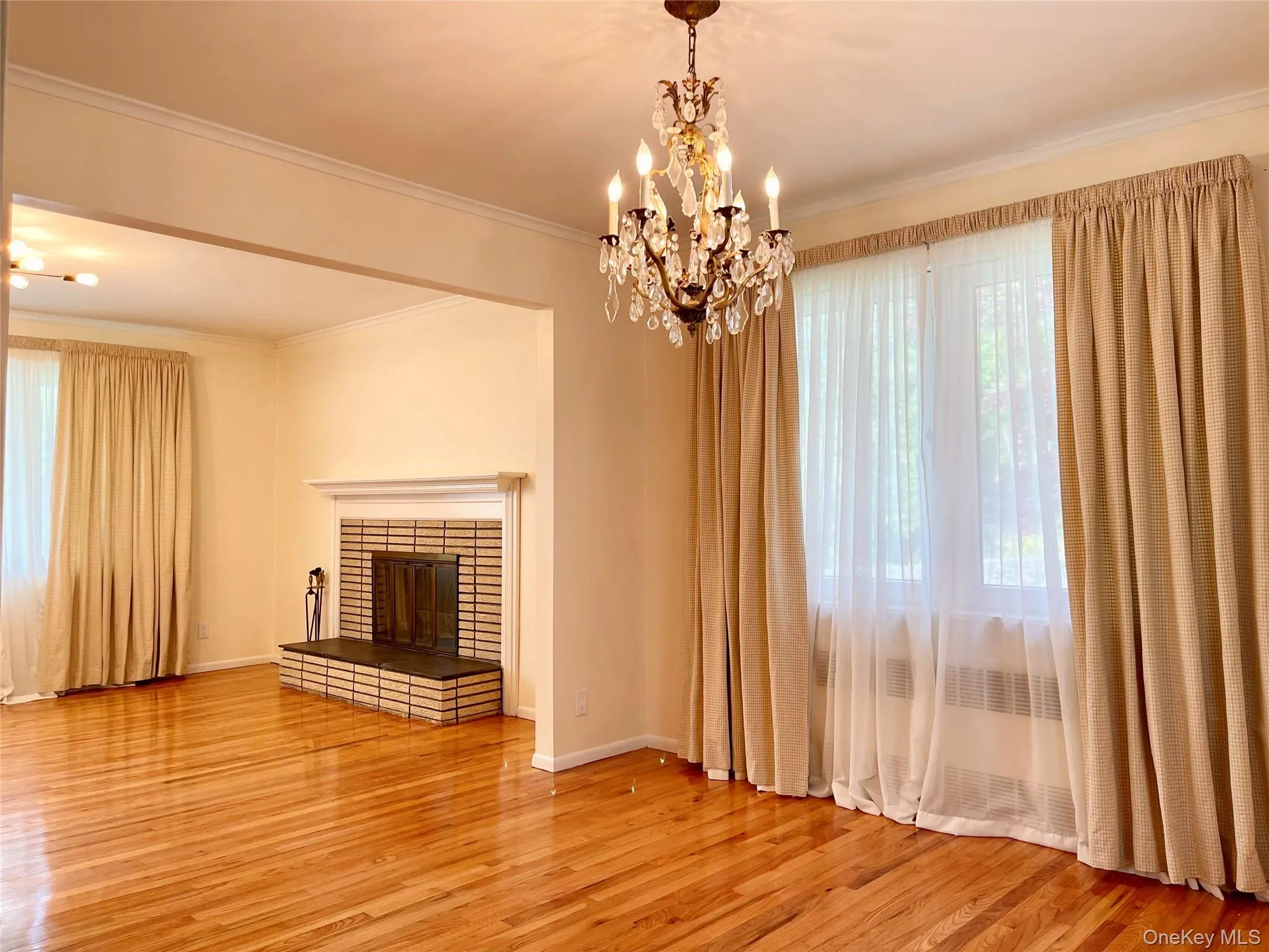 Unfurnished living room featuring a chandelier, radiator heating unit, crown molding, a brick fireplace, and light wood-style floors Unfurnished living room featuring a chandelier, radiator heating unit, crown molding, a brick fireplace, and light wood-style floors