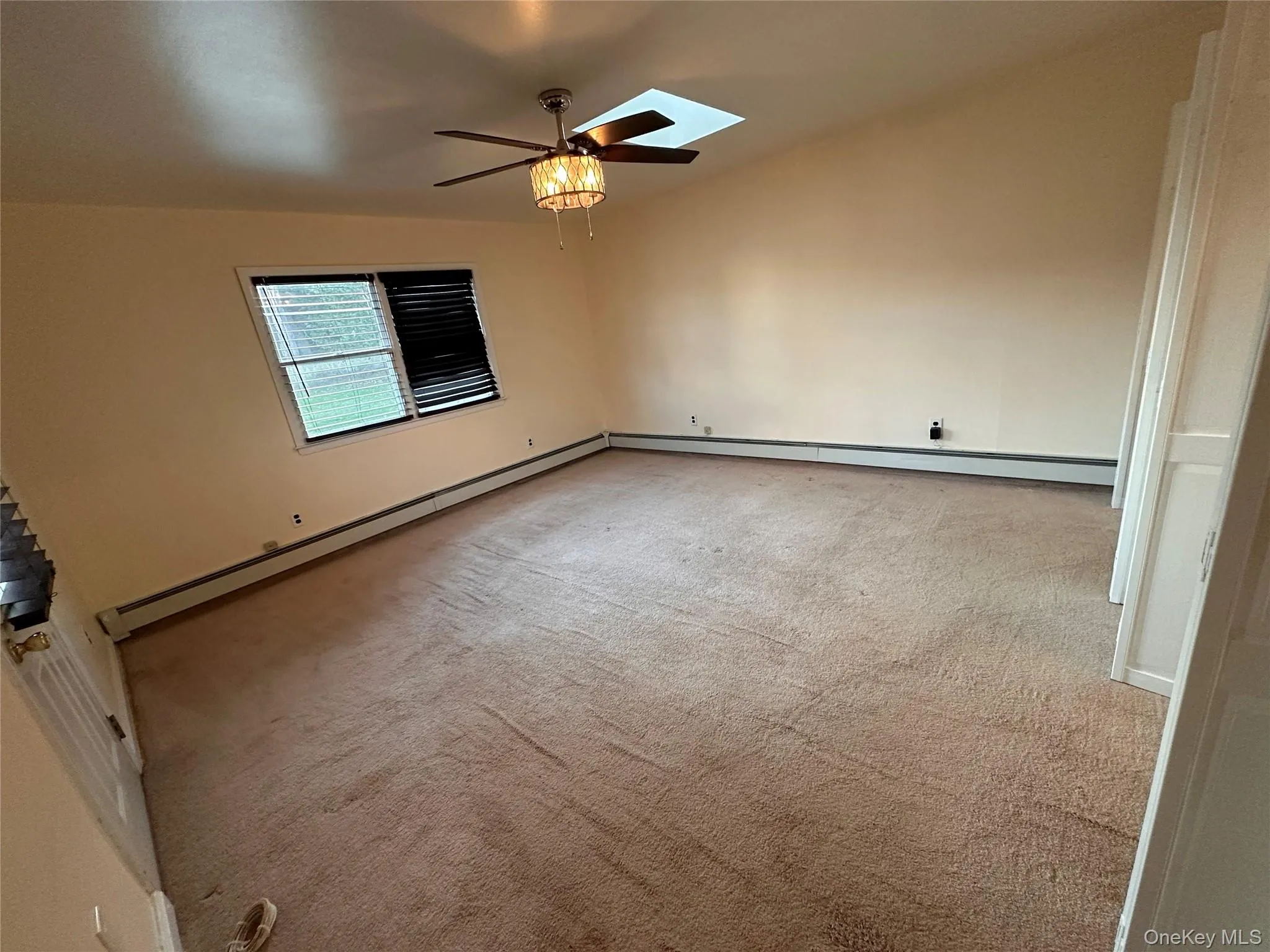 Empty room featuring a skylight, light colored carpet, a baseboard heating unit, and a ceiling fan Empty room featuring a skylight, light colored carpet, a baseboard heating unit, and a ceiling fan