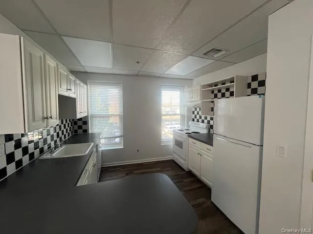 Kitchen featuring white appliances, white cabinets, dark countertops, a drop ceiling, and open shelves Kitchen featuring white appliances, white cabinets, dark countertops, a drop ceiling, and open shelves