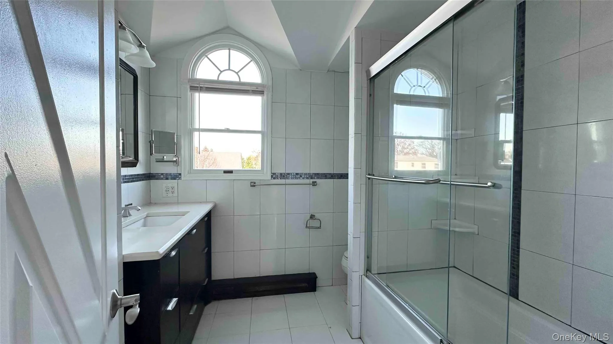 Bathroom featuring tile walls, vanity, light tile patterned flooring, bath / shower combo with glass door, and plenty of natural light Bathroom featuring tile walls, vanity, light tile patterned flooring, bath / shower combo with glass door, and plenty of natural light
