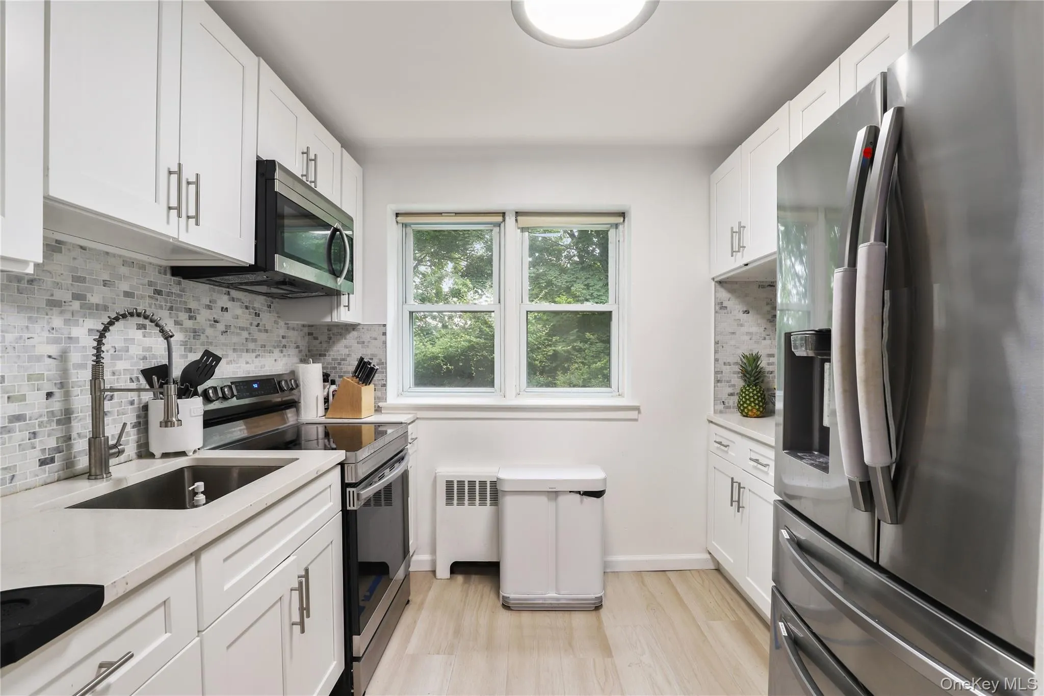 Kitchen featuring appliances with stainless steel finishes, radiator, light countertops, light wood-type flooring, and tasteful backsplash Kitchen featuring appliances with stainless steel finishes, radiator, light countertops, light wood-type flooring, and tasteful backsplash