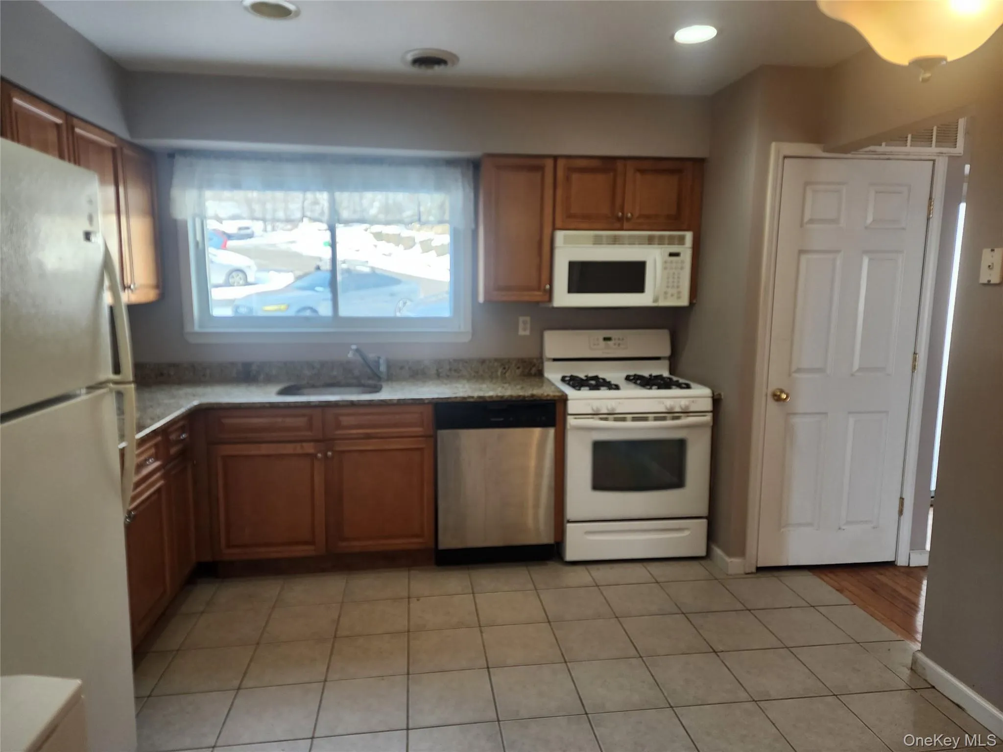 Kitchen with white appliances, brown cabinetry, light stone counters, light tile patterned floors, and recessed lighting Kitchen with white appliances, brown cabinetry, light stone counters, light tile patterned floors, and recessed lighting