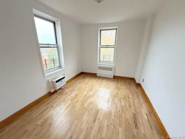 Unfurnished room featuring light wood-type flooring, radiator heating unit, and a wall unit AC Unfurnished room featuring light wood-type flooring, radiator heating unit, and a wall unit AC