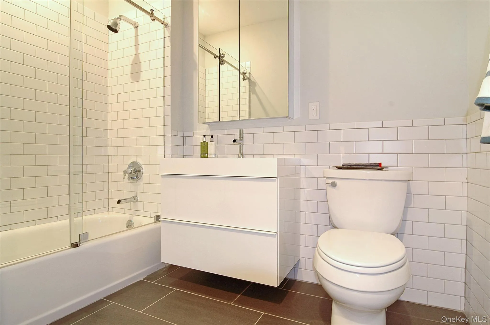 Full bathroom with tile walls, vanity, dark tile patterned floors, bath / shower combo with glass door, and a wainscoted wall Full bathroom with tile walls, vanity, dark tile patterned floors, bath / shower combo with glass door, and a wainscoted wall