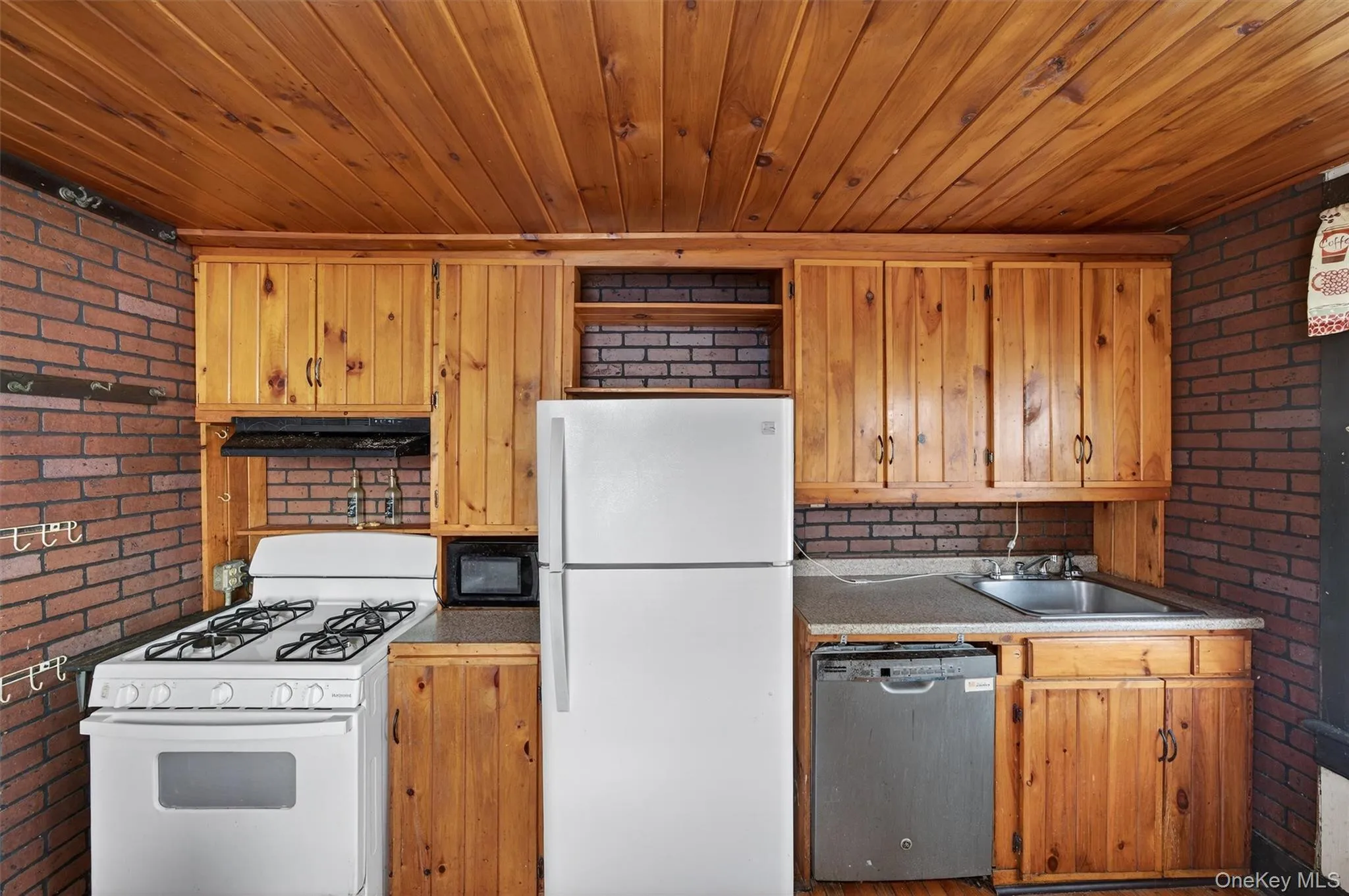 Kitchen with brick wall, white appliances, wooden ceiling, brown cabinets, and under cabinet range hood Kitchen with brick wall, white appliances, wooden ceiling, brown cabinets, and under cabinet range hood