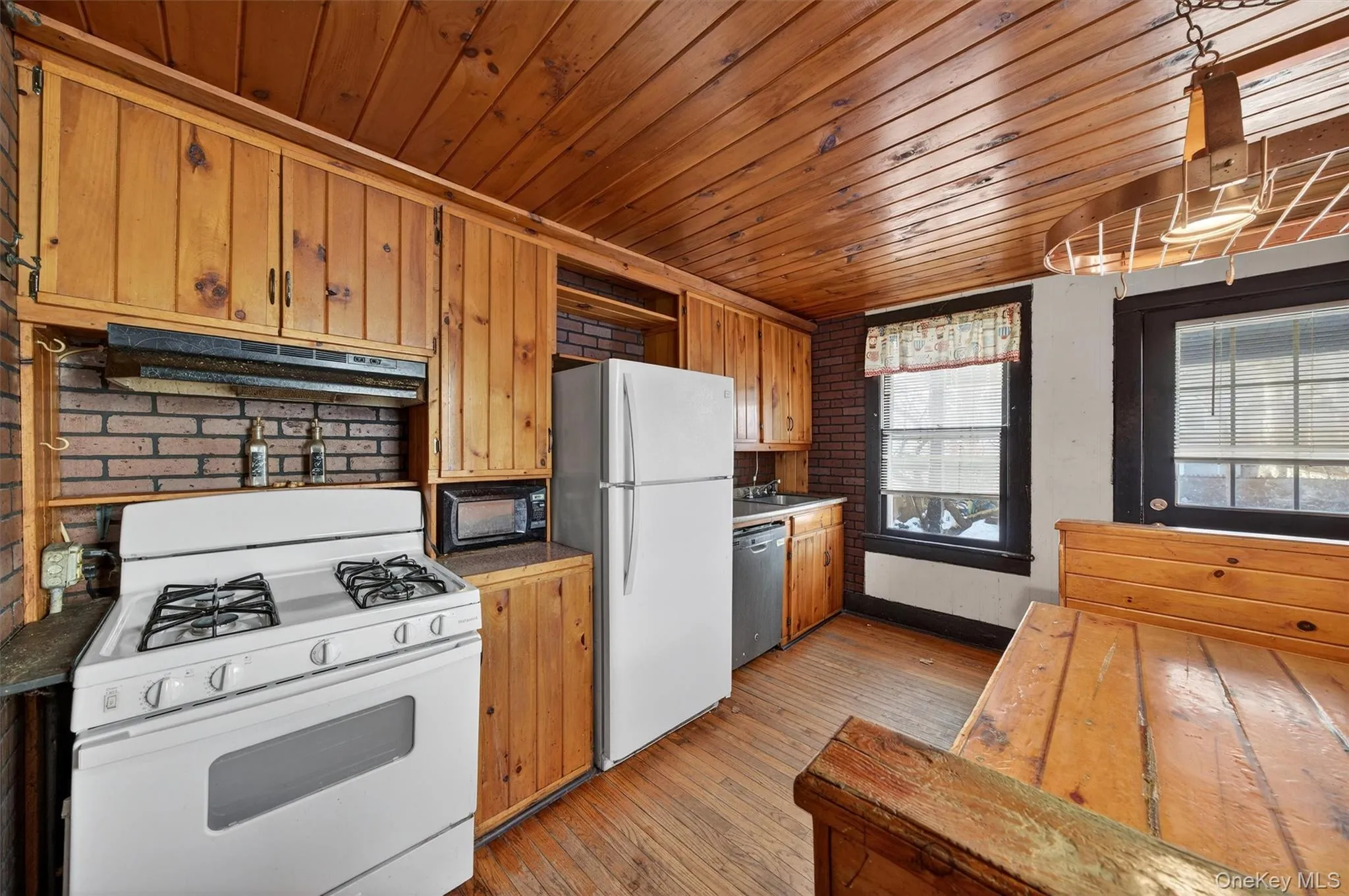 Kitchen featuring white appliances, brick wall, brown cabinets, under cabinet range hood, and wooden ceiling Kitchen featuring white appliances, brick wall, brown cabinets, under cabinet range hood, and wooden ceiling