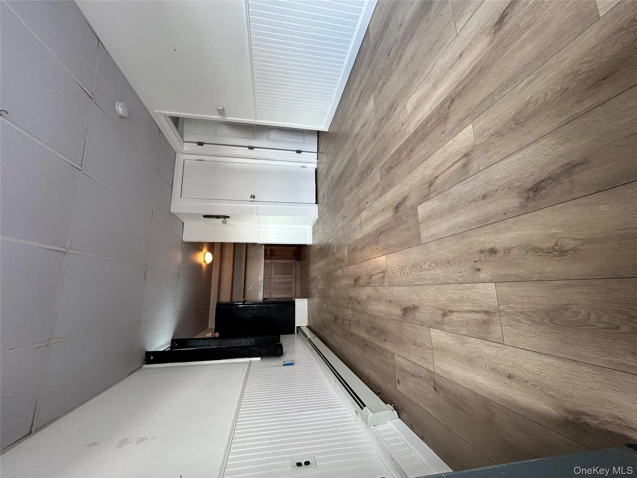 Unfurnished living room featuring a baseboard heating unit, light wood-type flooring, a drop ceiling, and wainscoting Unfurnished living room featuring a baseboard heating unit, light wood-type flooring, a drop ceiling, and wainscoting