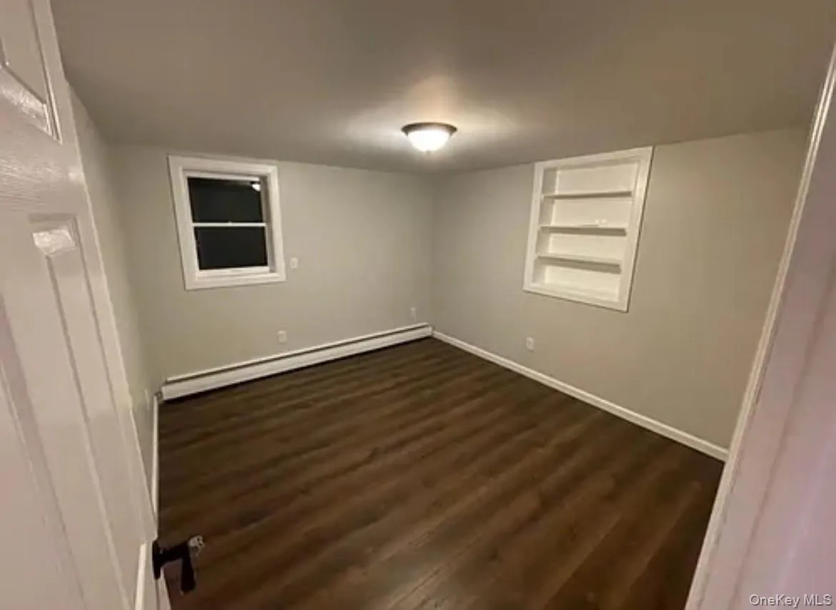 Empty room featuring built in shelves, dark wood-style floors, and baseboard heating Empty room featuring built in shelves, dark wood-style floors, and baseboard heating