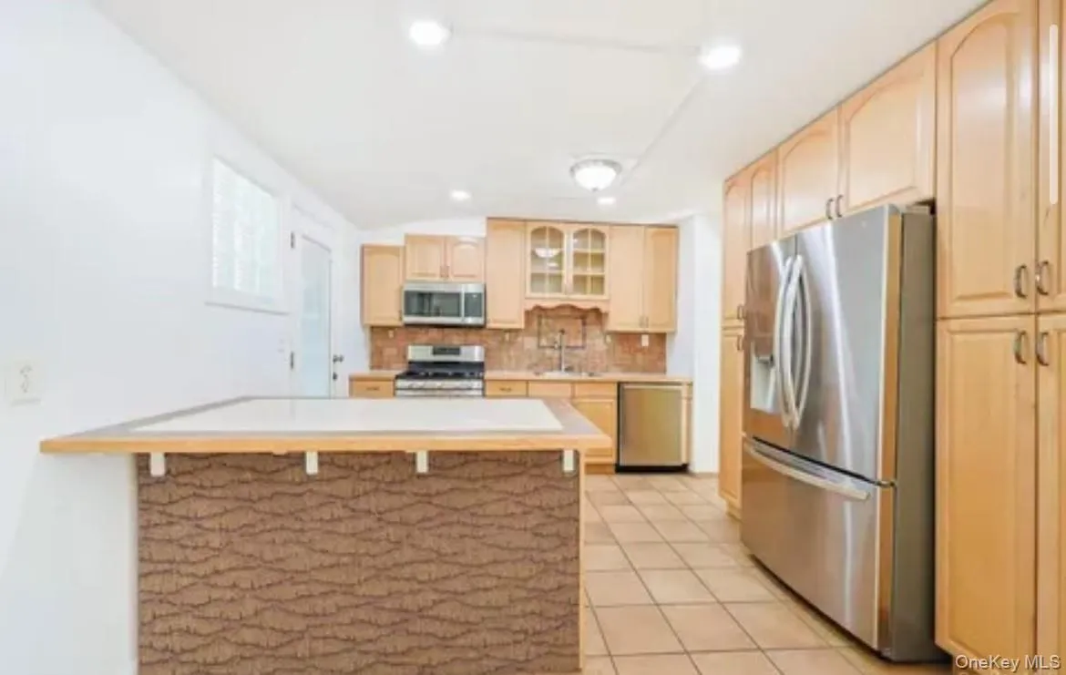 Kitchen featuring light countertops, appliances with stainless steel finishes, glass insert cabinets, a kitchen bar, and backsplash Kitchen featuring light countertops, appliances with stainless steel finishes, glass insert cabinets, a kitchen bar, and backsplash
