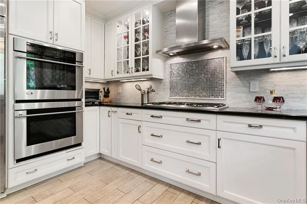 Kitchen featuring appliances with stainless steel finishes, wall chimney exhaust hood, white cabinets, glass insert cabinets, and backsplash Kitchen featuring appliances with stainless steel finishes, wall chimney exhaust hood, white cabinets, glass insert cabinets, and backsplash