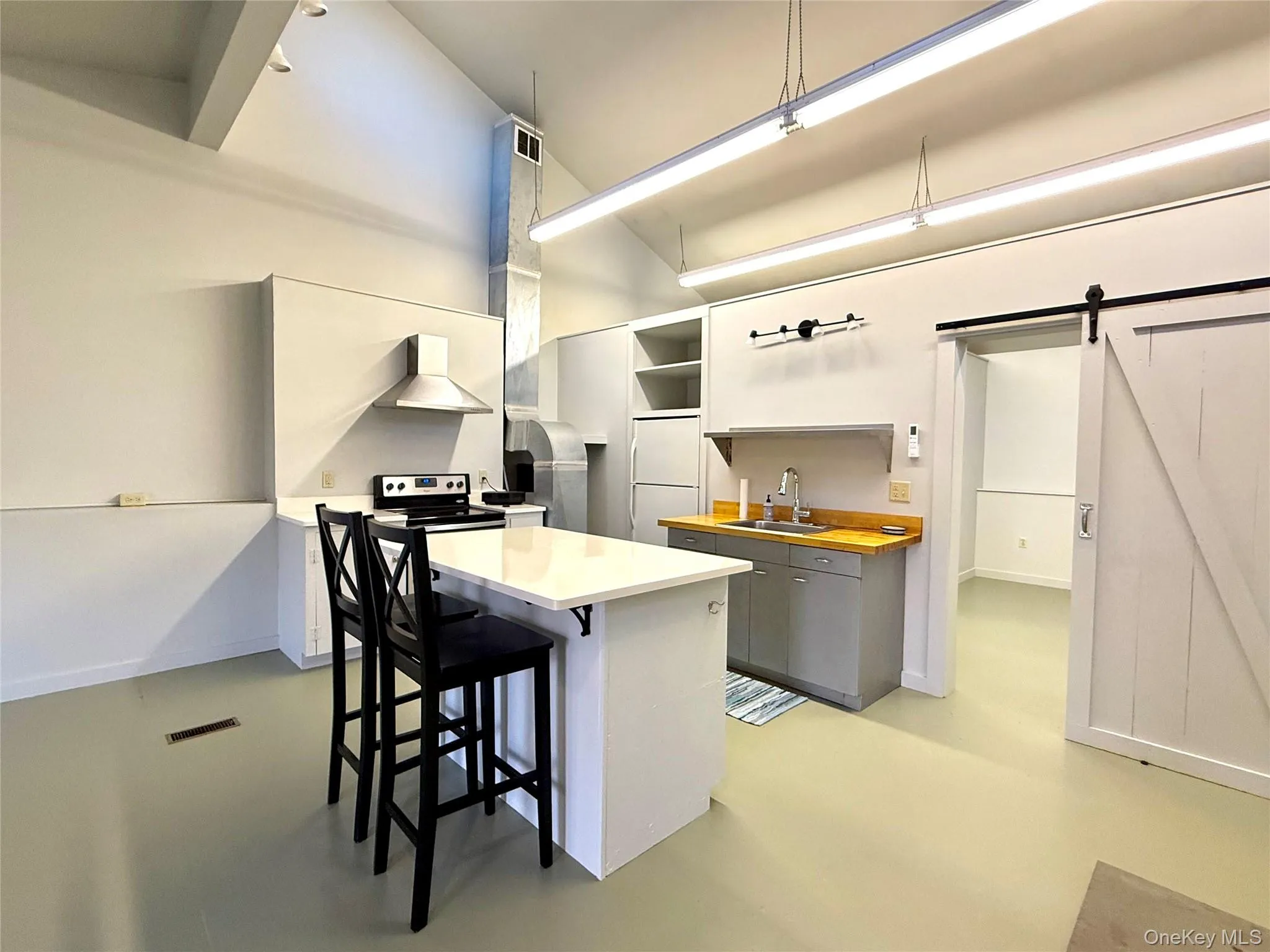 Kitchen with concrete flooring, stainless steel range with electric stovetop, wall chimney exhaust hood, a barn door, and a breakfast bar Kitchen with concrete flooring, stainless steel range with electric stovetop, wall chimney exhaust hood, a barn door, and a breakfast bar