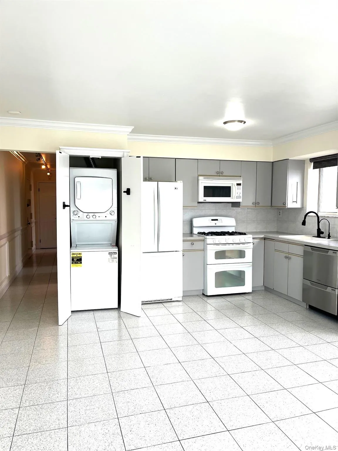 Kitchen with gray cabinets, white appliances, ornamental molding, stacked washer and clothes dryer, and light countertops Kitchen with gray cabinets, white appliances, ornamental molding, stacked washer and clothes dryer, and light countertops