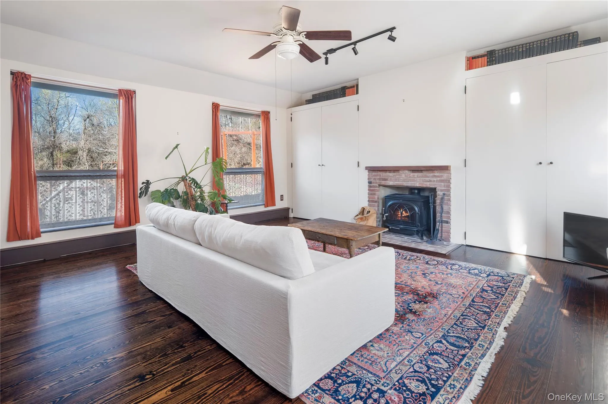 additional living area in back of home, or possible primary bedroom, featuring massive floor to ceiling windows that flood the space with natural light, track lighting, a ceiling fan, hardwood flooring and cast iron Jotul wood-burning stove insert in fireplace additional living area in back of home, or possible primary bedroom, featuring massive floor to ceiling windows that flood the space with natural light, track lighting, a ceiling fan, hardwood flooring and cast iron Jotul wood-burning stove insert in fireplace