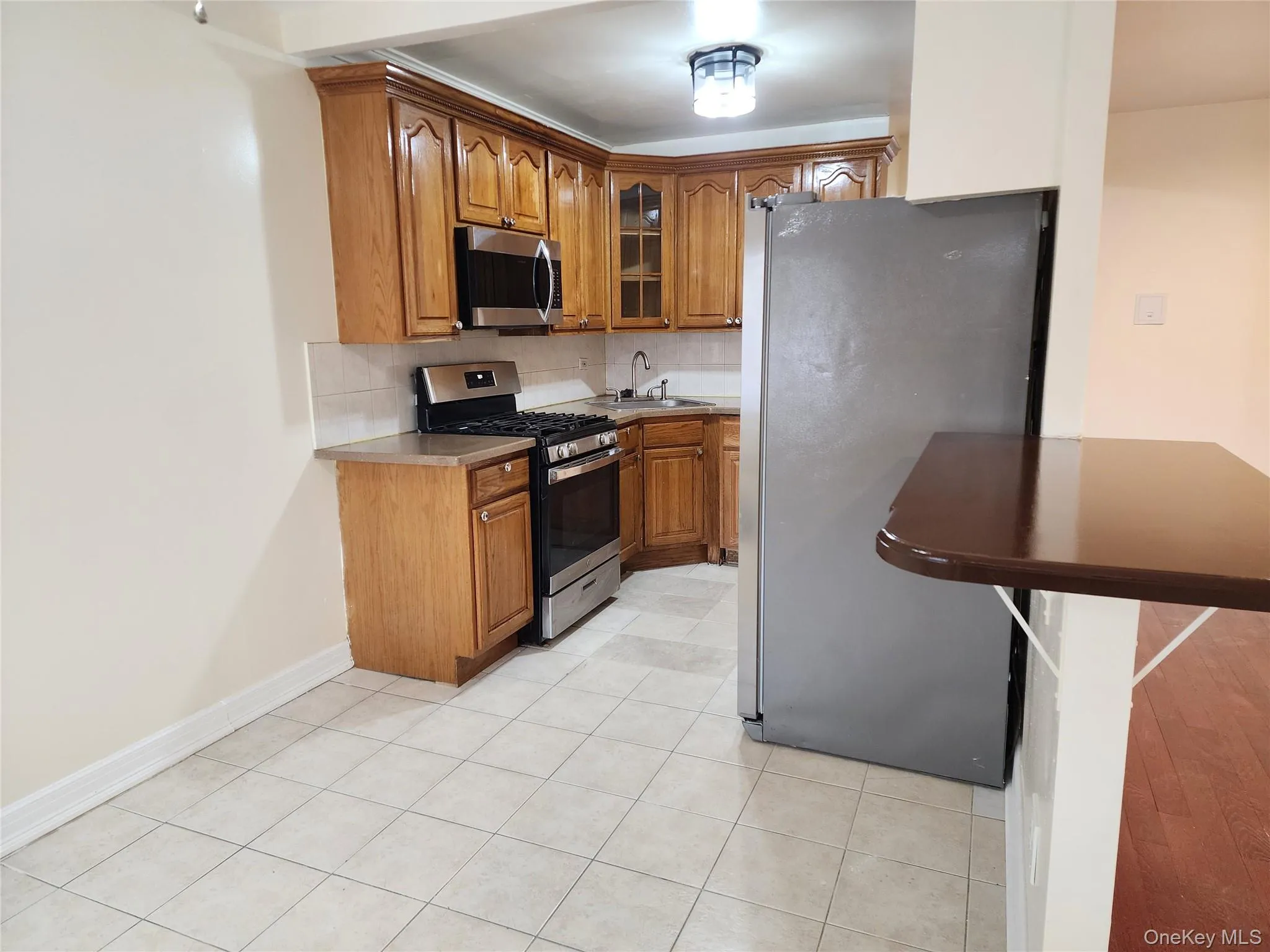Kitchen featuring appliances with stainless steel finishes, brown cabinetry, tasteful backsplash, glass insert cabinets, and light tile patterned flooring Kitchen featuring appliances with stainless steel finishes, brown cabinetry, tasteful backsplash, glass insert cabinets, and light tile patterned flooring