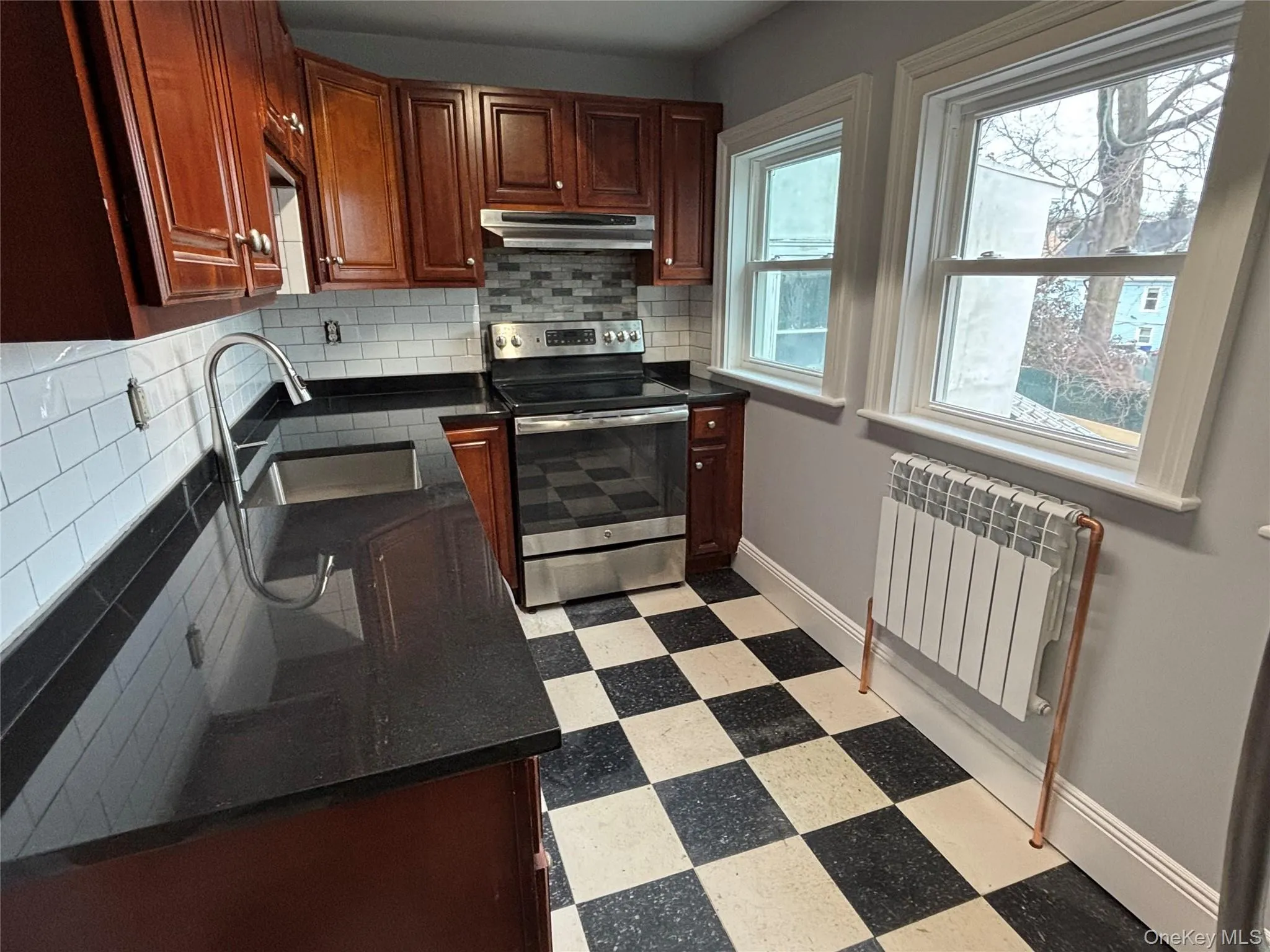 Kitchen featuring stainless steel range with electric stovetop, radiator heating unit, checkerboard floors floors, dark granite counter tops, and decorative subway tile backsplash Kitchen featuring stainless steel range with electric stovetop, radiator heating unit, checkerboard floors floors, dark granite counter tops, and decorative subway tile backsplash