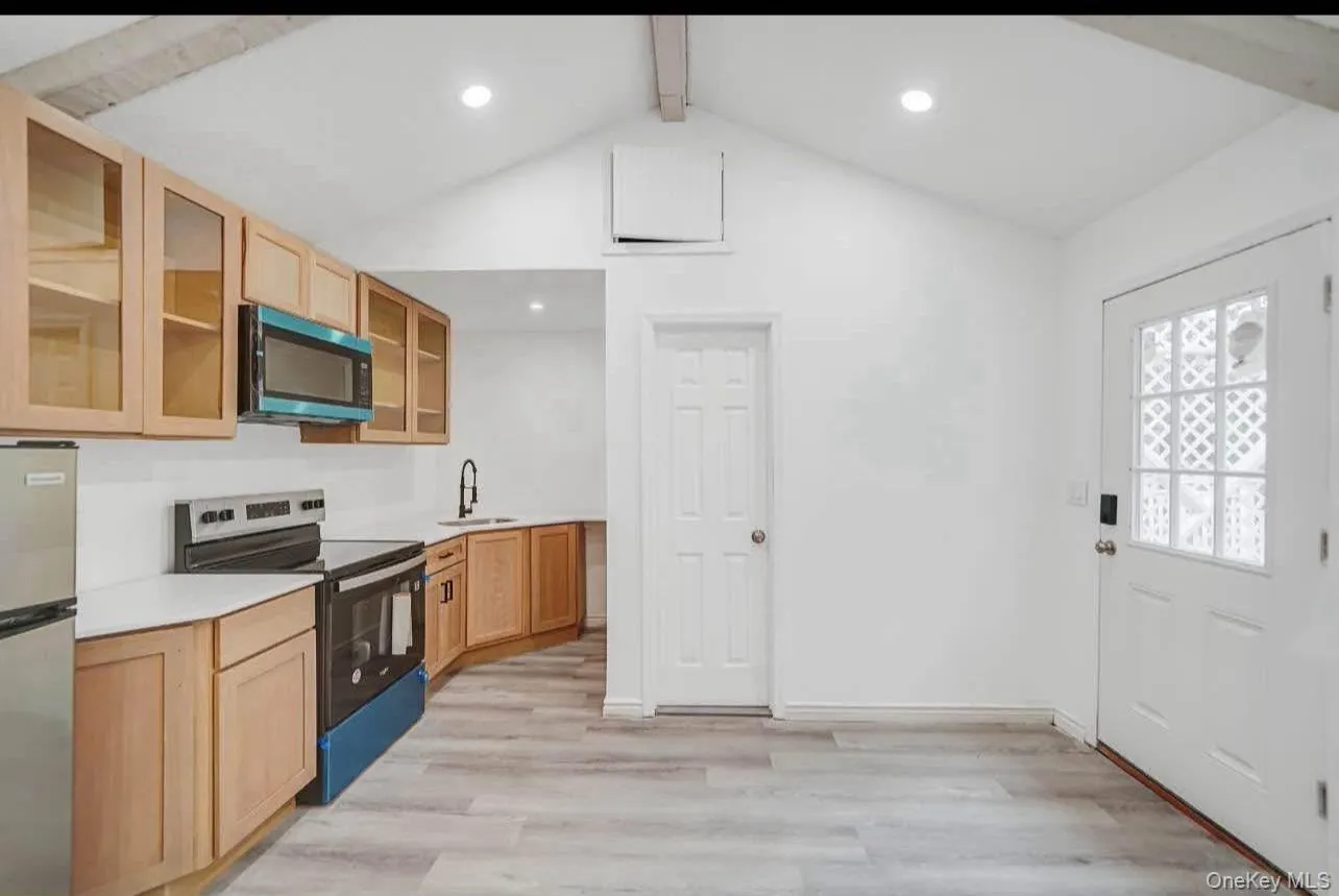 Kitchen featuring stainless steel appliances, light countertops, light wood-type flooring, glass insert cabinets, and light brown cabinets Kitchen featuring stainless steel appliances, light countertops, light wood-type flooring, glass insert cabinets, and light brown cabinets
