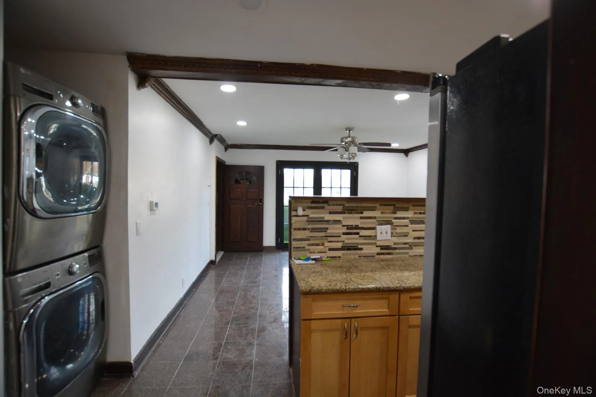 Kitchen featuring ornamental molding, brown cabinetry, decorative backsplash, stacked washer / dryer, and light stone countertops Kitchen featuring ornamental molding, brown cabinetry, decorative backsplash, stacked washer / dryer, and light stone countertops