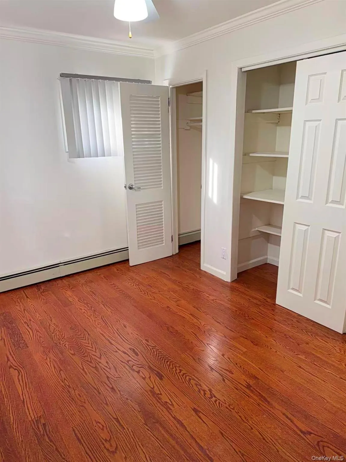 Unfurnished bedroom with crown molding, multiple closets, baseboard heating, wood finished floors, and ceiling fan Unfurnished bedroom with crown molding, multiple closets, baseboard heating, wood finished floors, and ceiling fan