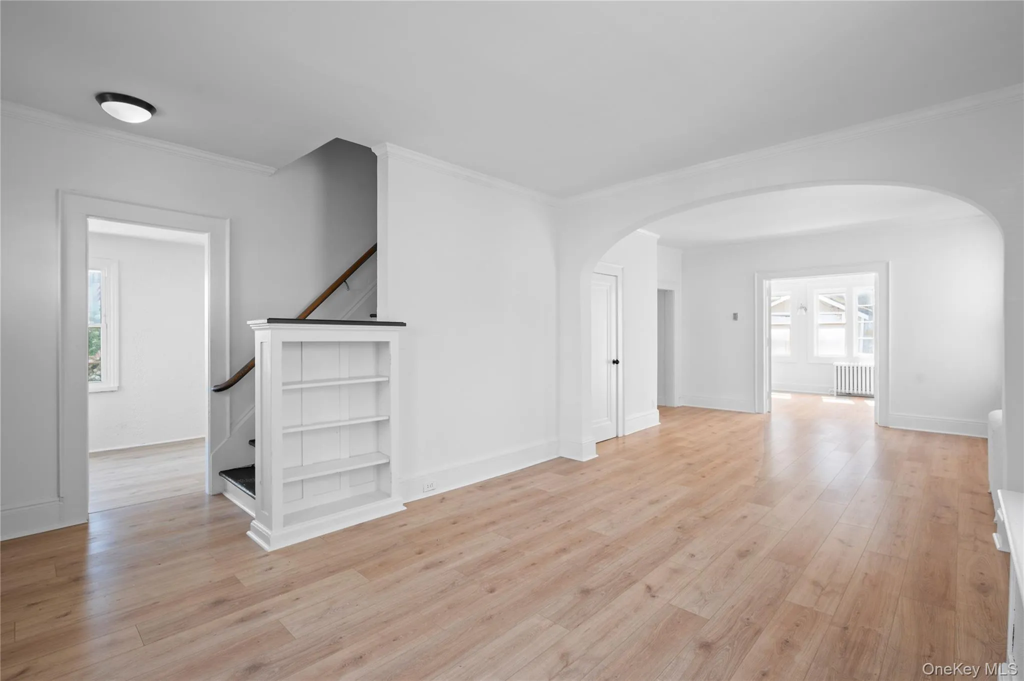 Unfurnished living room featuring light wood-style flooring, arched walkways, crown molding, radiator heating unit, and stairway Unfurnished living room featuring light wood-style flooring, arched walkways, crown molding, radiator heating unit, and stairway