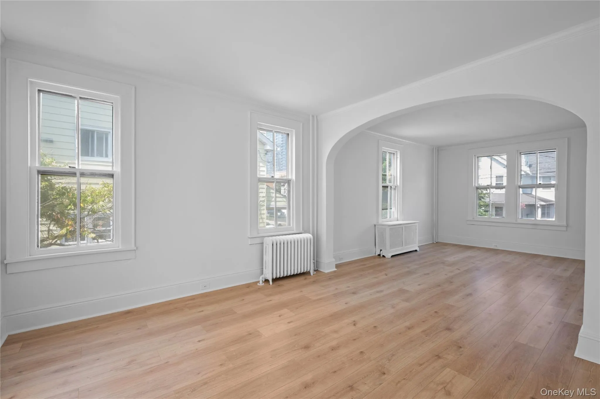 Empty room featuring arched walkways, radiator heating unit, light wood finished floors, healthy amount of natural light, and ornamental molding Empty room featuring arched walkways, radiator heating unit, light wood finished floors, healthy amount of natural light, and ornamental molding