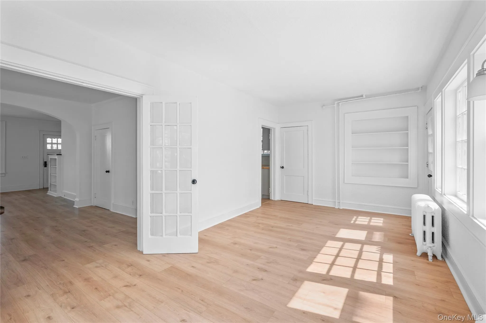 Unfurnished room featuring built in shelves, radiator heating unit, light wood-type flooring, and arched walkways Unfurnished room featuring built in shelves, radiator heating unit, light wood-type flooring, and arched walkways