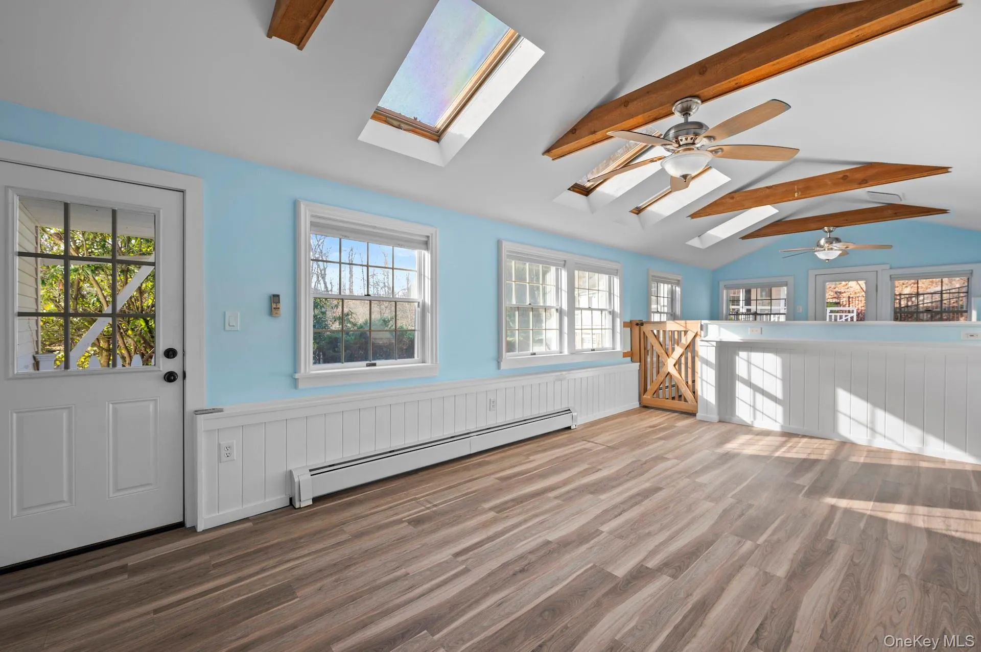 Unfurnished living room with a wainscoted wall, baseboard heating, wood finished floors, a skylight, and plenty of natural light Unfurnished living room with a wainscoted wall, baseboard heating, wood finished floors, a skylight, and plenty of natural light