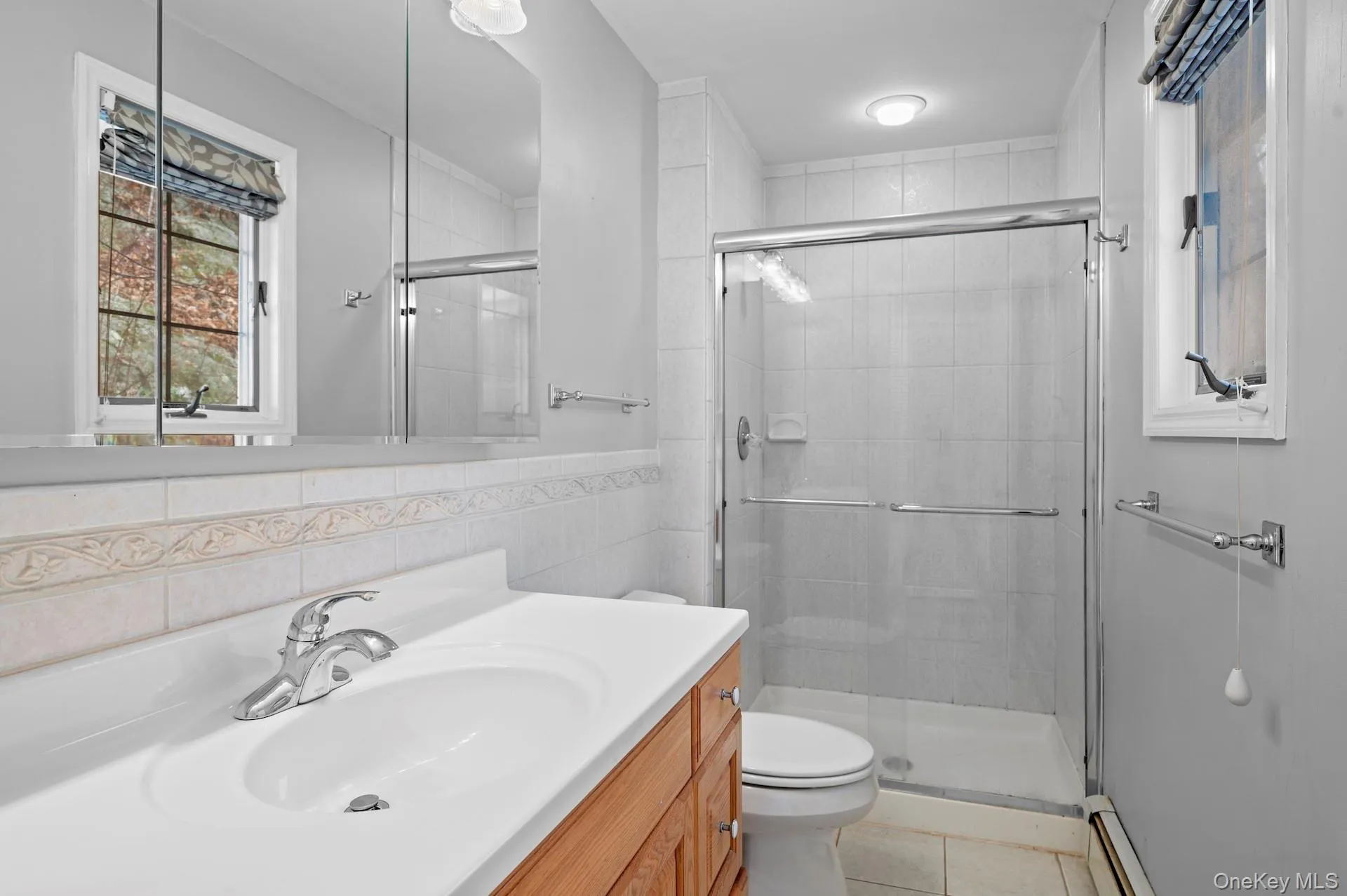 Full bath featuring vanity, tile walls, a stall shower, a baseboard heating unit, and light tile patterned floors Full bath featuring vanity, tile walls, a stall shower, a baseboard heating unit, and light tile patterned floors