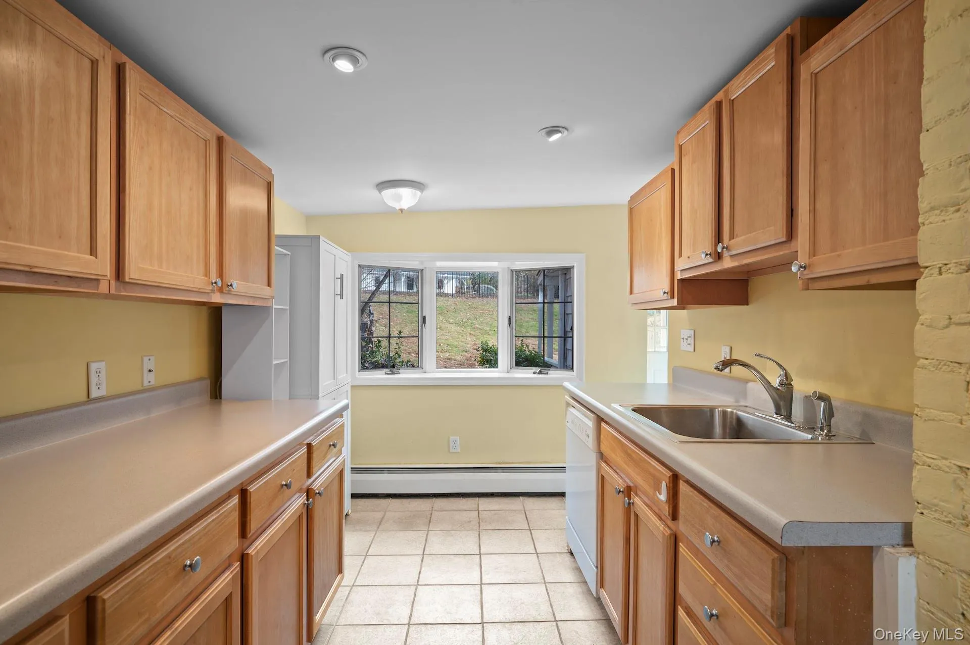Kitchen featuring a baseboard radiator, light countertops, white dishwasher, light tile patterned floors, and brown cabinets Kitchen featuring a baseboard radiator, light countertops, white dishwasher, light tile patterned floors, and brown cabinets