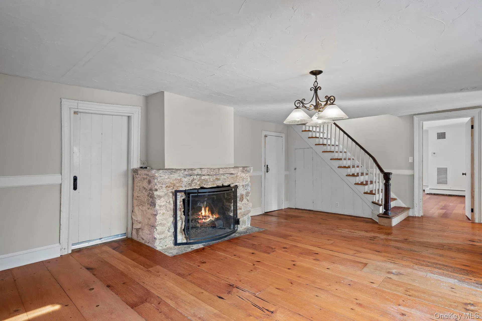 Unfurnished living room featuring a stone fireplace, stairway, wood-type flooring, a chandelier, and baseboard heating Unfurnished living room featuring a stone fireplace, stairway, wood-type flooring, a chandelier, and baseboard heating