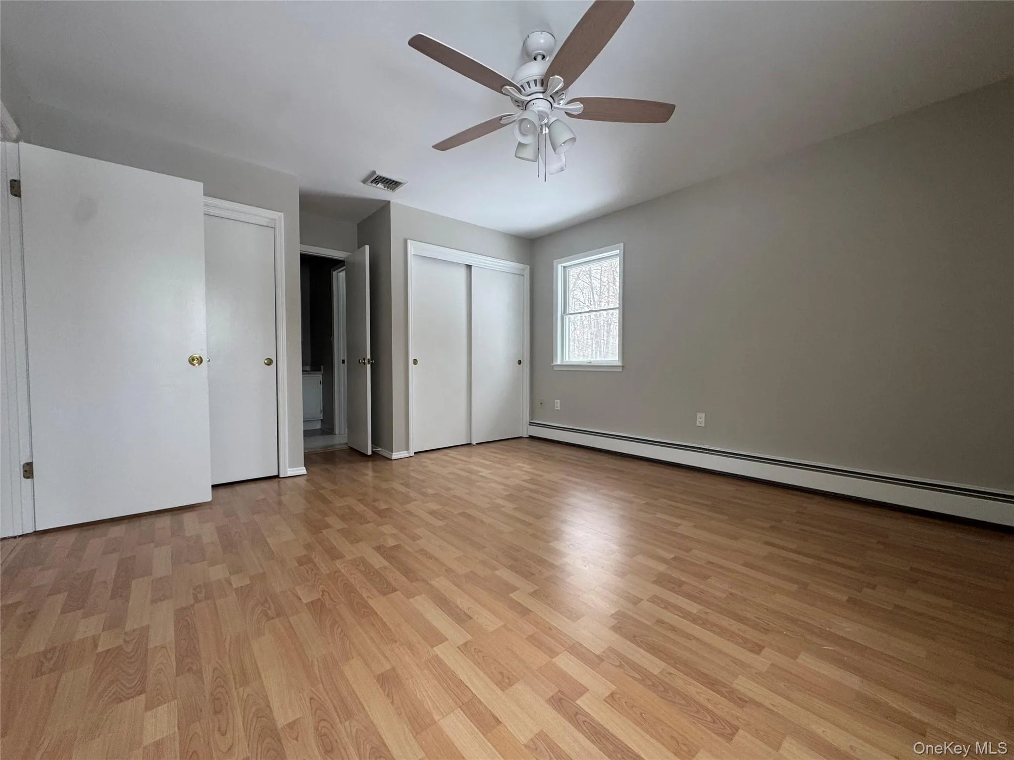 Unfurnished bedroom featuring light wood-type flooring, a baseboard heating unit, a ceiling fan, and multiple closets Unfurnished bedroom featuring light wood-type flooring, a baseboard heating unit, a ceiling fan, and multiple closets
