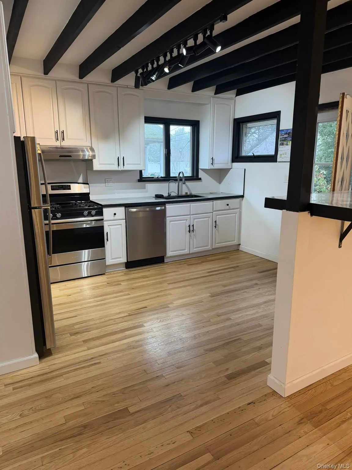 Kitchen featuring stainless steel appliances, white cabinets, beam ceiling, light wood finished floors, and dark countertops Kitchen featuring stainless steel appliances, white cabinets, beam ceiling, light wood finished floors, and dark countertops