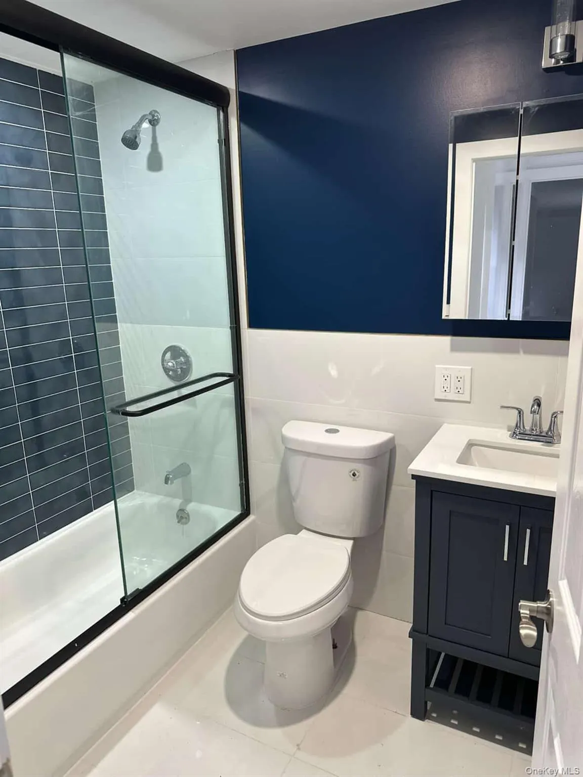 Bathroom featuring vanity, bath / shower combo with glass door, tile walls, light tile patterned floors, and a wainscoted wall Bathroom featuring vanity, bath / shower combo with glass door, tile walls, light tile patterned floors, and a wainscoted wall