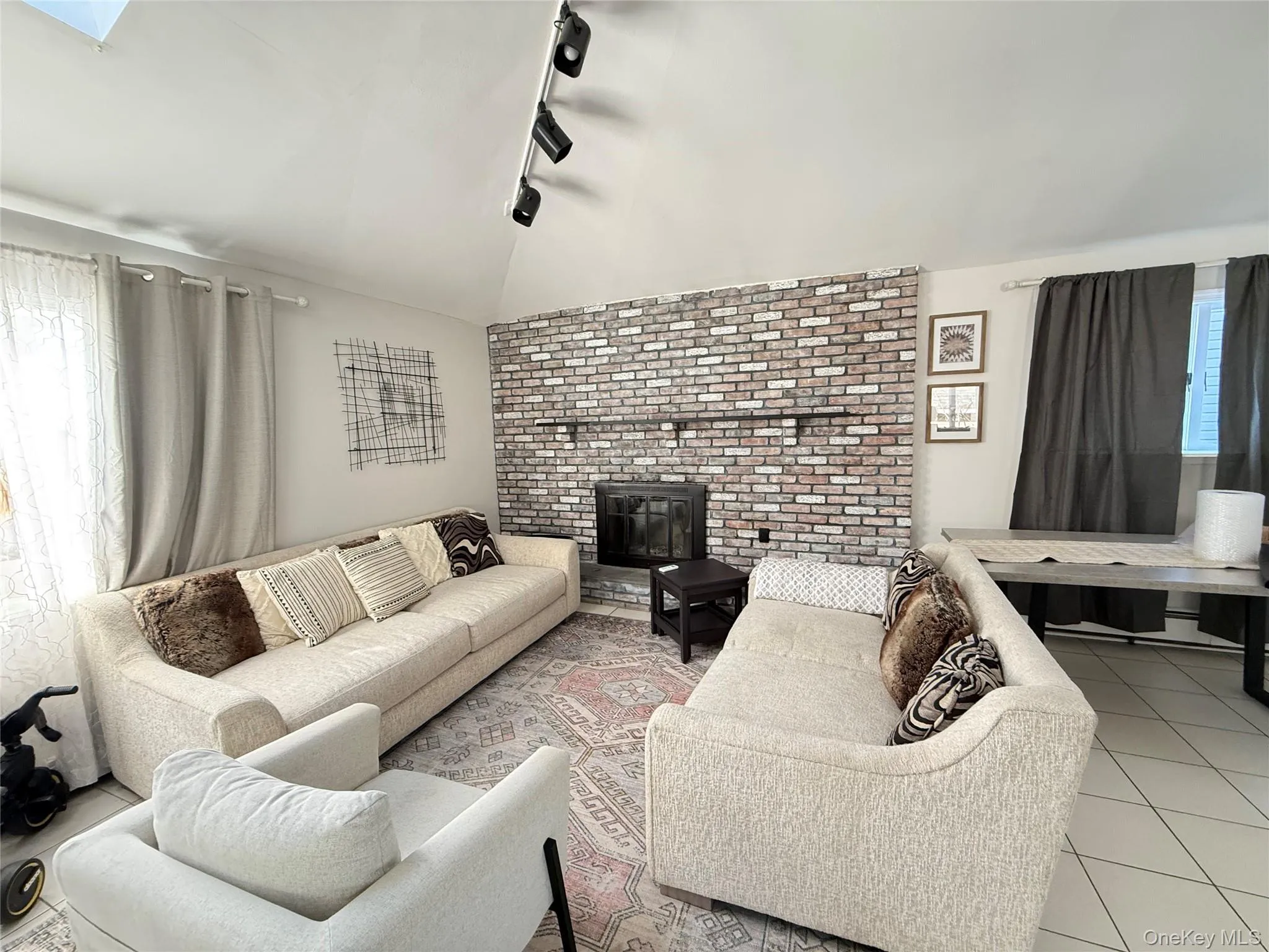 Tiled living area featuring vaulted ceiling, rail lighting, a brick fireplace, and a skylight Tiled living area featuring vaulted ceiling, rail lighting, a brick fireplace, and a skylight
