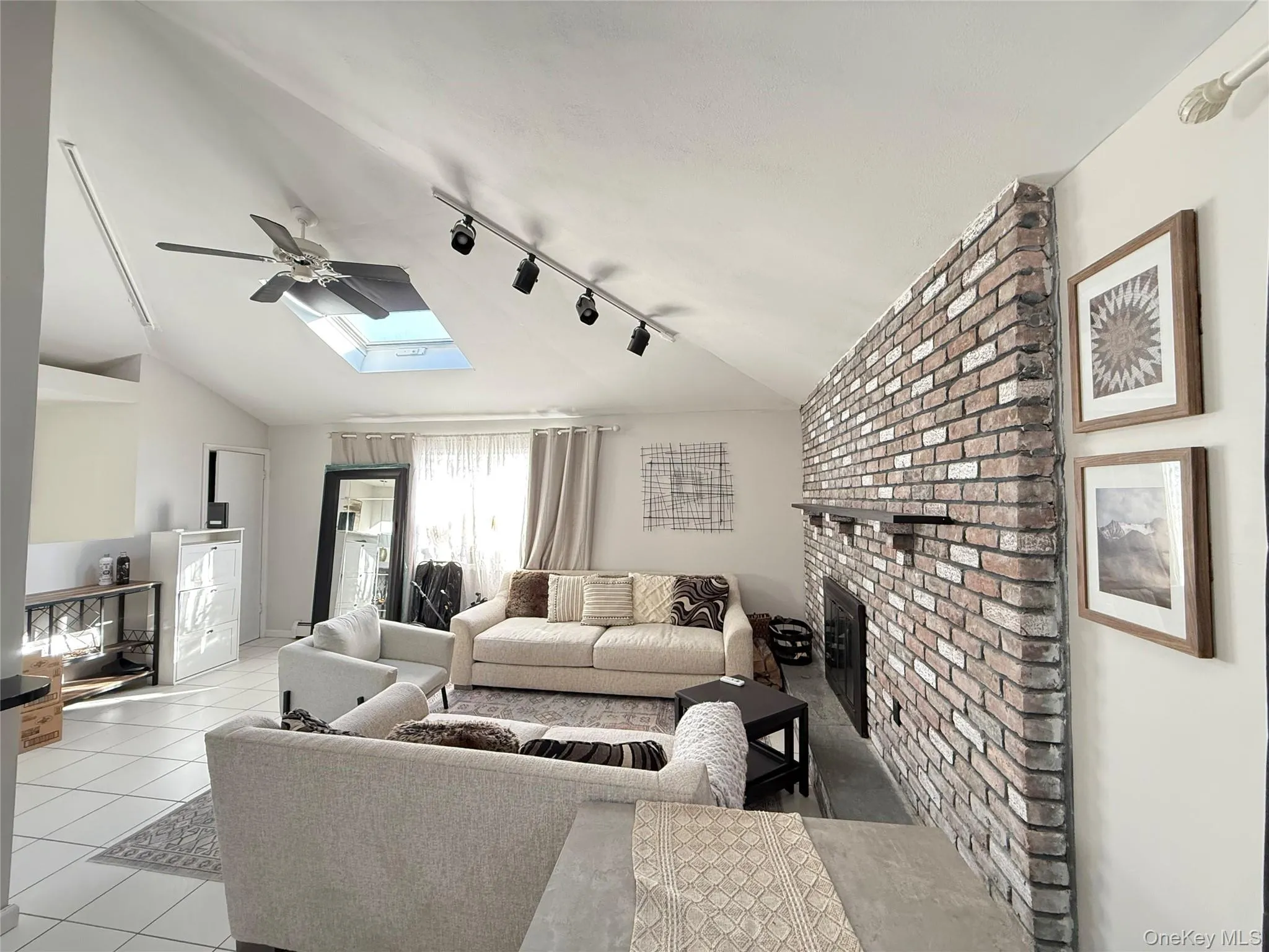 Living room featuring a brick fireplace, vaulted ceiling, a skylight, rail lighting, and light tile patterned flooring Living room featuring a brick fireplace, vaulted ceiling, a skylight, rail lighting, and light tile patterned flooring