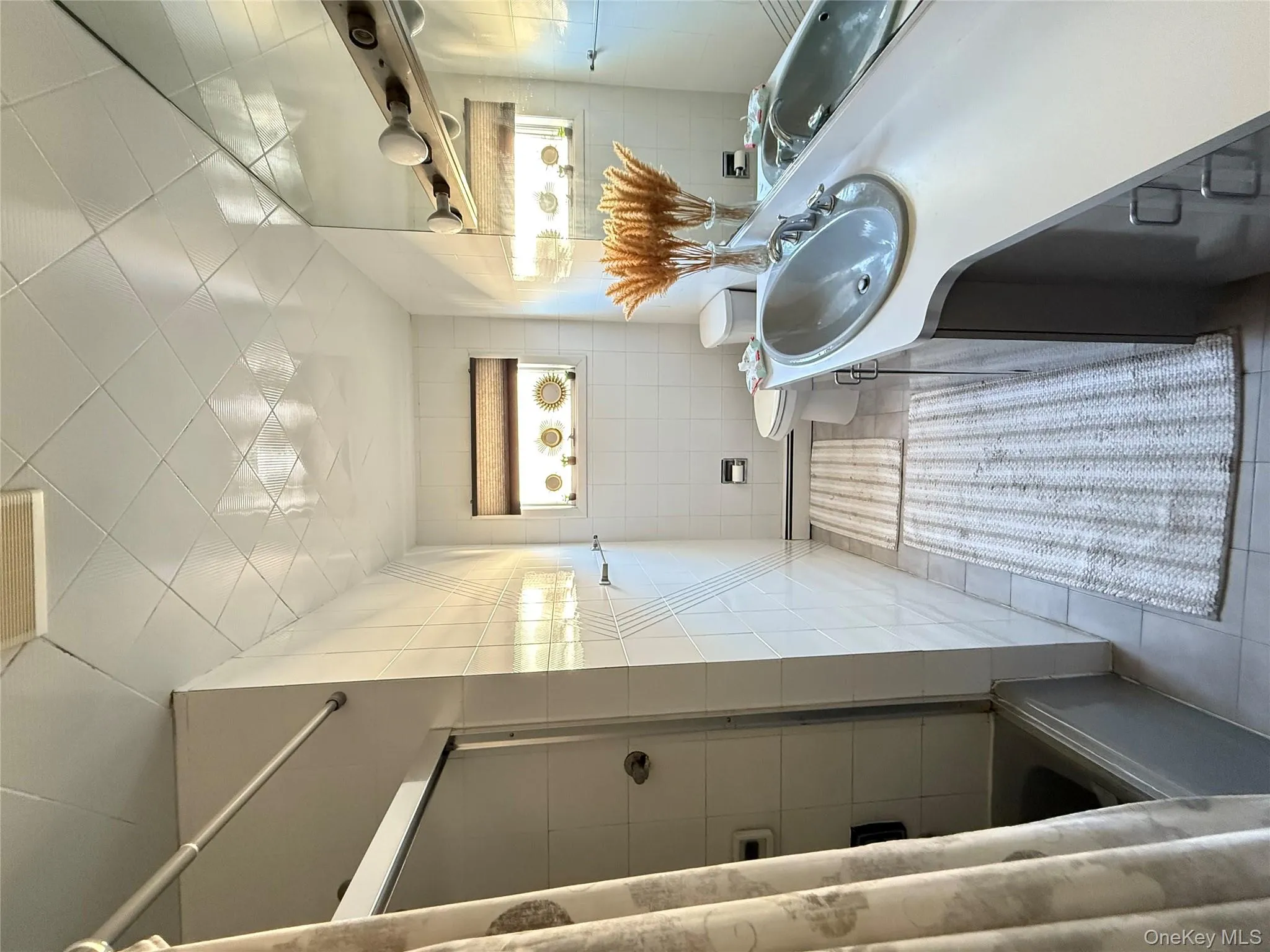 Full bath with light tile patterned flooring, bath / shower combo with glass door, vanity, tile walls, and a baseboard heating unit Full bath with light tile patterned flooring, bath / shower combo with glass door, vanity, tile walls, and a baseboard heating unit