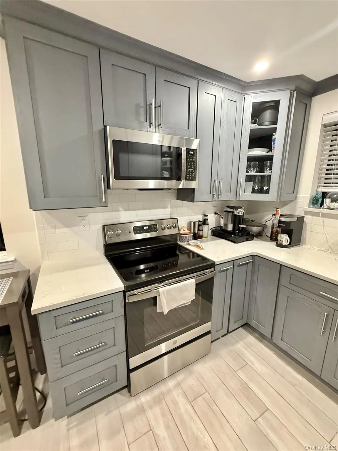 Kitchen with gray cabinets, tasteful backsplash, stainless steel appliances, glass insert cabinets, and light stone counters Kitchen with gray cabinets, tasteful backsplash, stainless steel appliances, glass insert cabinets, and light stone counters
