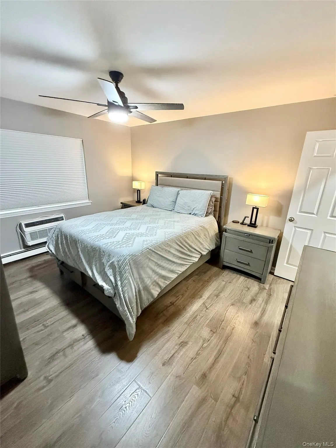 Bedroom featuring baseboard heating, ceiling fan, light wood-style floors, and a wall mounted air conditioner Bedroom featuring baseboard heating, ceiling fan, light wood-style floors, and a wall mounted air conditioner