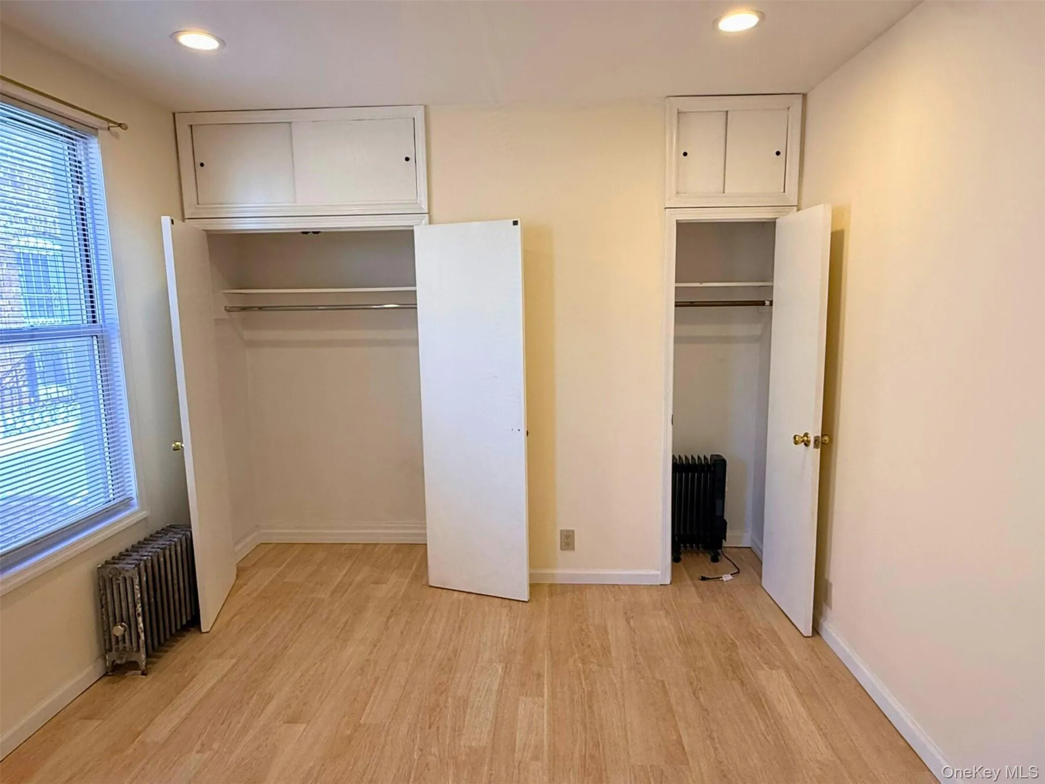 Unfurnished bedroom featuring radiator heating unit, light wood finished floors, recessed lighting, and multiple closets Unfurnished bedroom featuring radiator heating unit, light wood finished floors, recessed lighting, and multiple closets