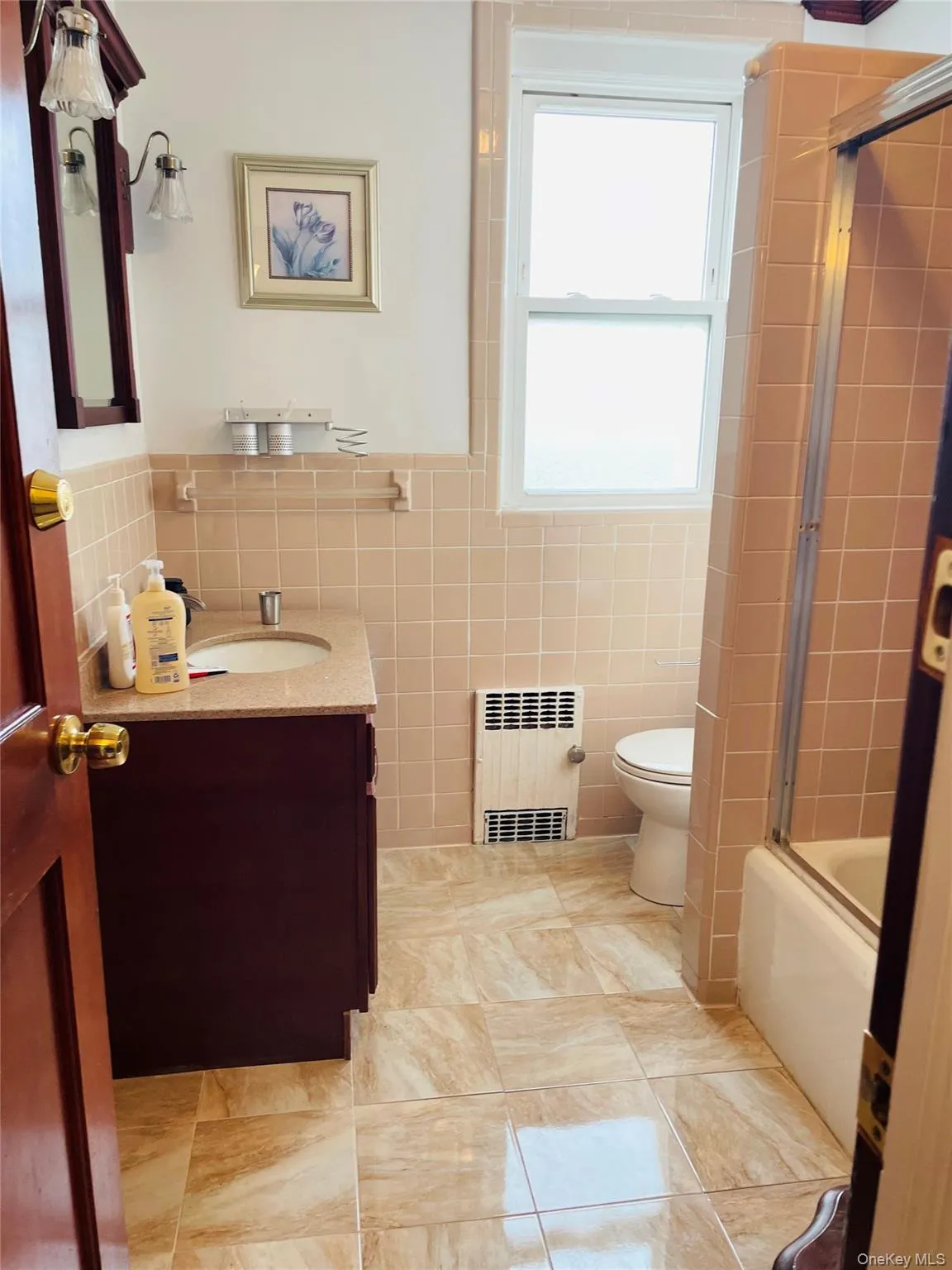 Full bathroom featuring vanity, tile walls, radiator, bath / shower combo with glass door, and wainscoting Full bathroom featuring vanity, tile walls, radiator, bath / shower combo with glass door, and wainscoting
