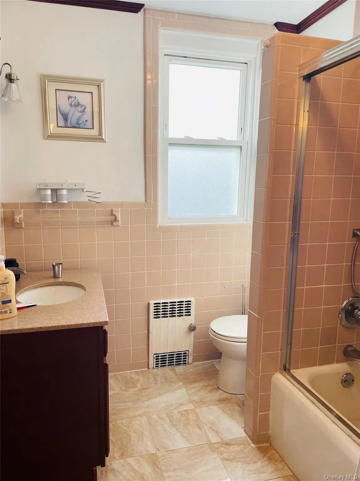 Full bath featuring tile walls, vanity, bath / shower combo with glass door, and radiator heating unit Full bath featuring tile walls, vanity, bath / shower combo with glass door, and radiator heating unit