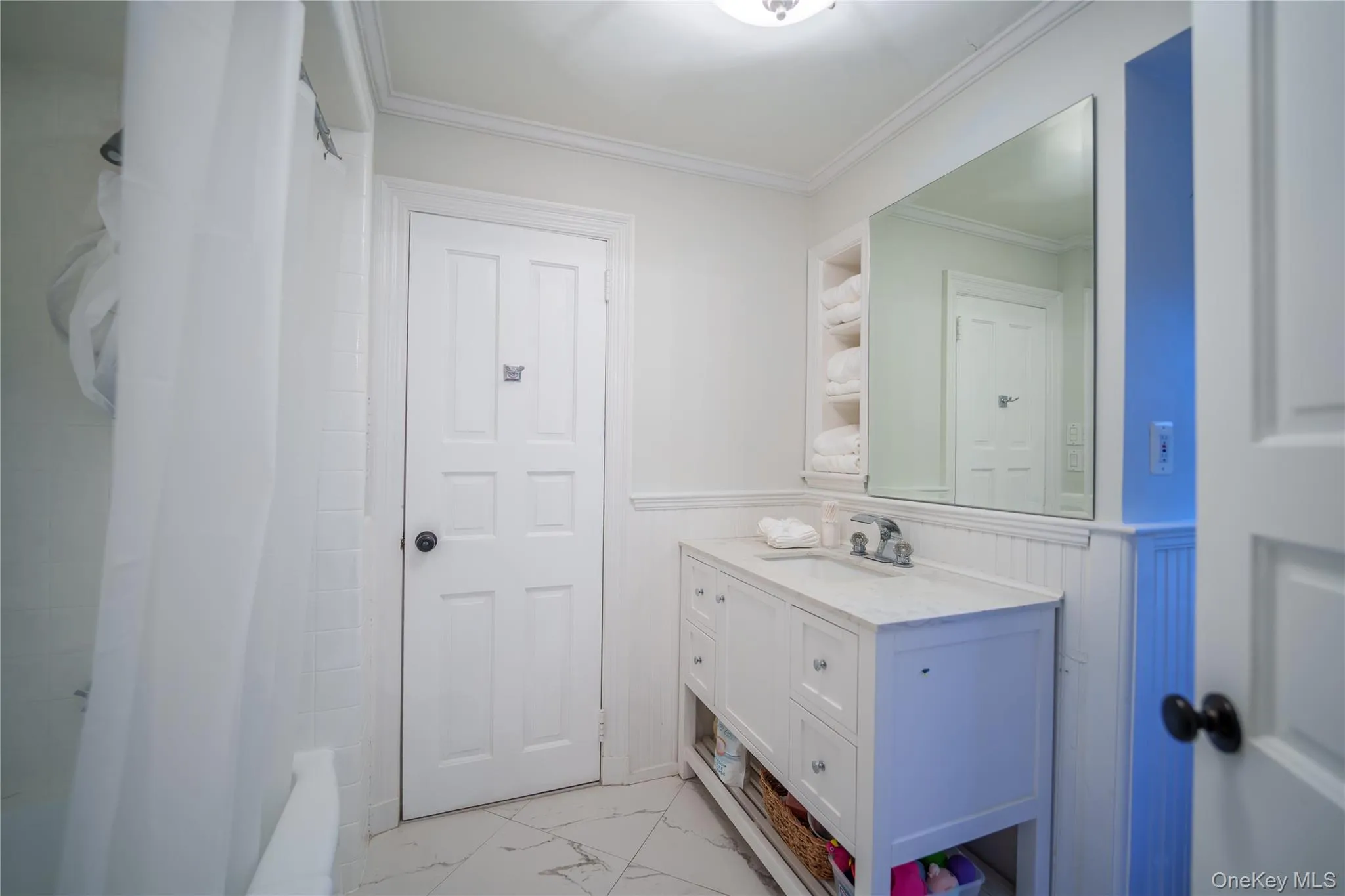 Full bath featuring a wainscoted wall, large vanity, ornamental molding, light marble finish floors, and shower / bathtub combination with curtain Full bath featuring a wainscoted wall, large vanity, ornamental molding, light marble finish floors, and shower / bathtub combination with curtain