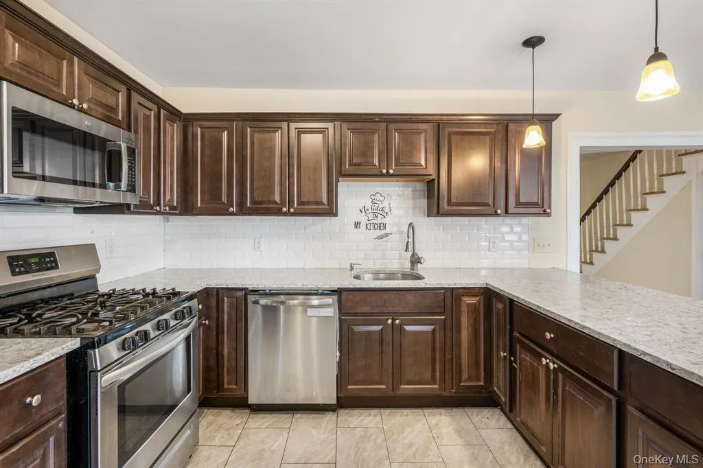 Kitchen with appliances with stainless steel finishes, dark brown cabinets, backsplash, hanging light fixtures, and light stone countertops Kitchen with appliances with stainless steel finishes, dark brown cabinets, backsplash, hanging light fixtures, and light stone countertops