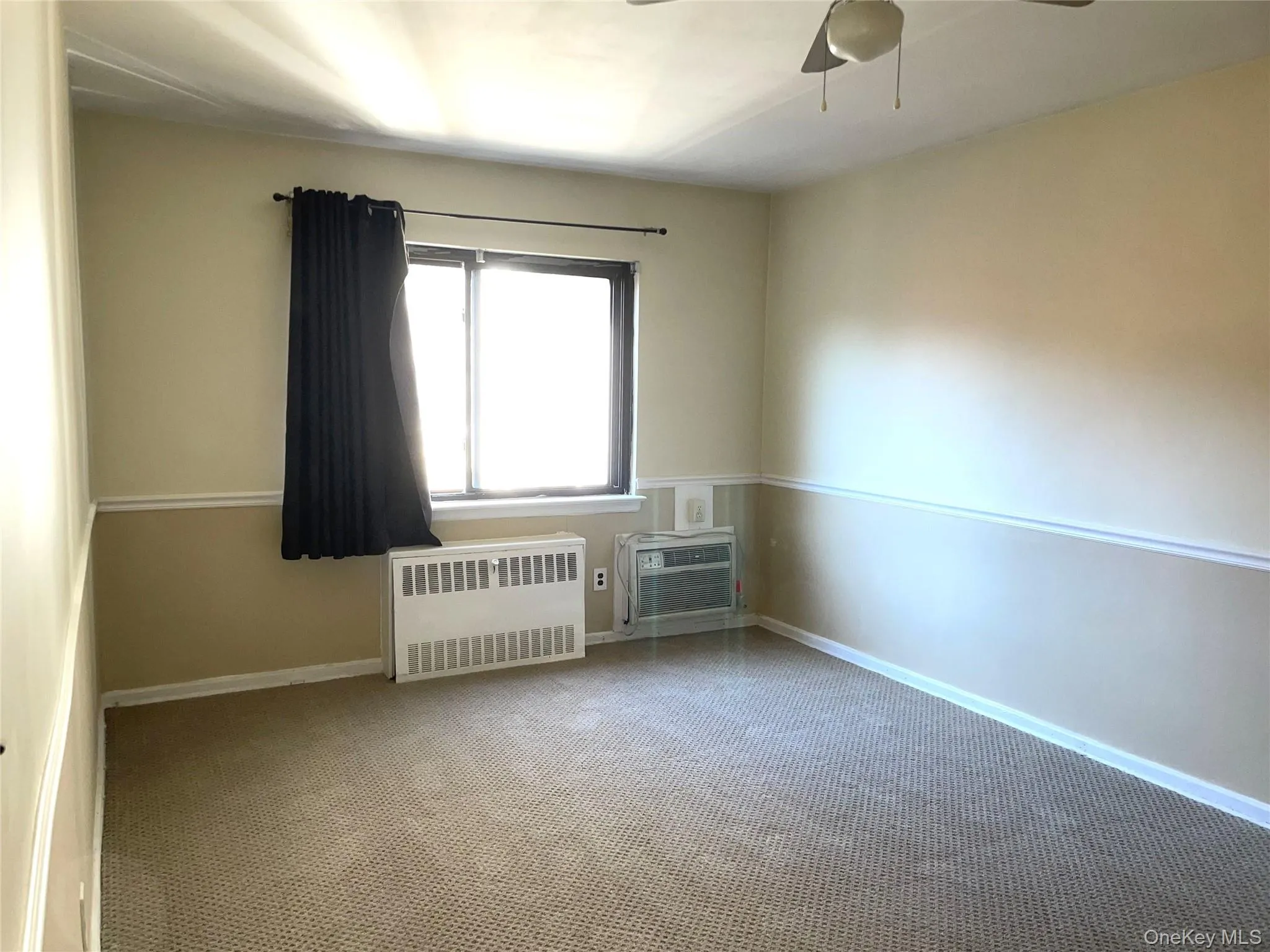 Spare room featuring carpet, radiator heating unit, a wall mounted air conditioner, and ceiling fan Spare room featuring carpet, radiator heating unit, a wall mounted air conditioner, and ceiling fan